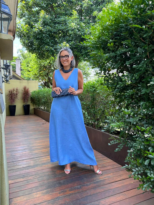 Mulher com vestido longo de linho azul, sandálias e bolsa, em varanda com plantas EZILDINHA