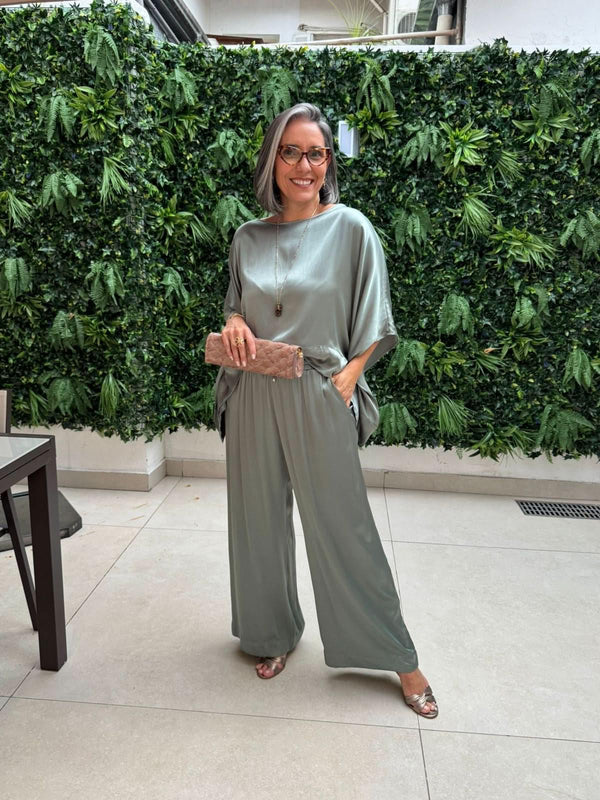 Mulher com cabelo grisalho usando conjunto de linho verde claro, segurando clutch nude, em ambiente com parede de folhagens EZILDINHA