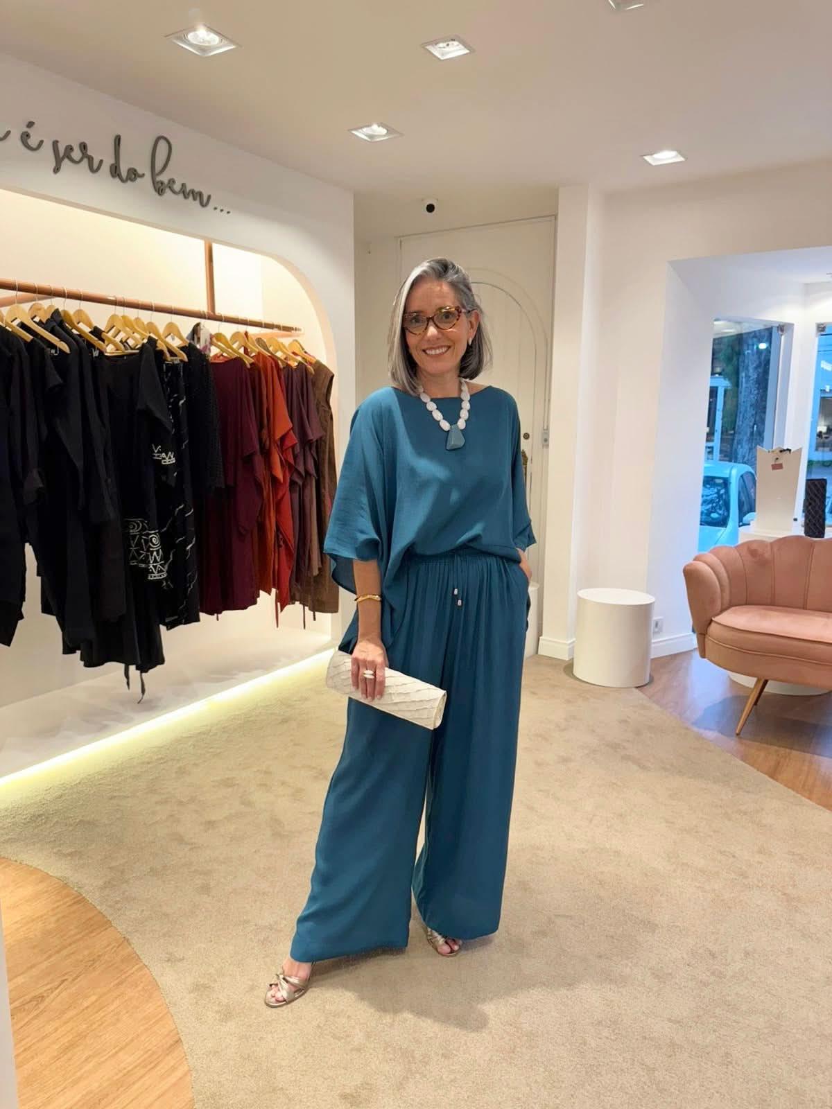 Mulher com cabelo grisalho usando conjunto azul elegante de tecido leve e colar moderno em loja de roupas sofisticada EZILDINHA