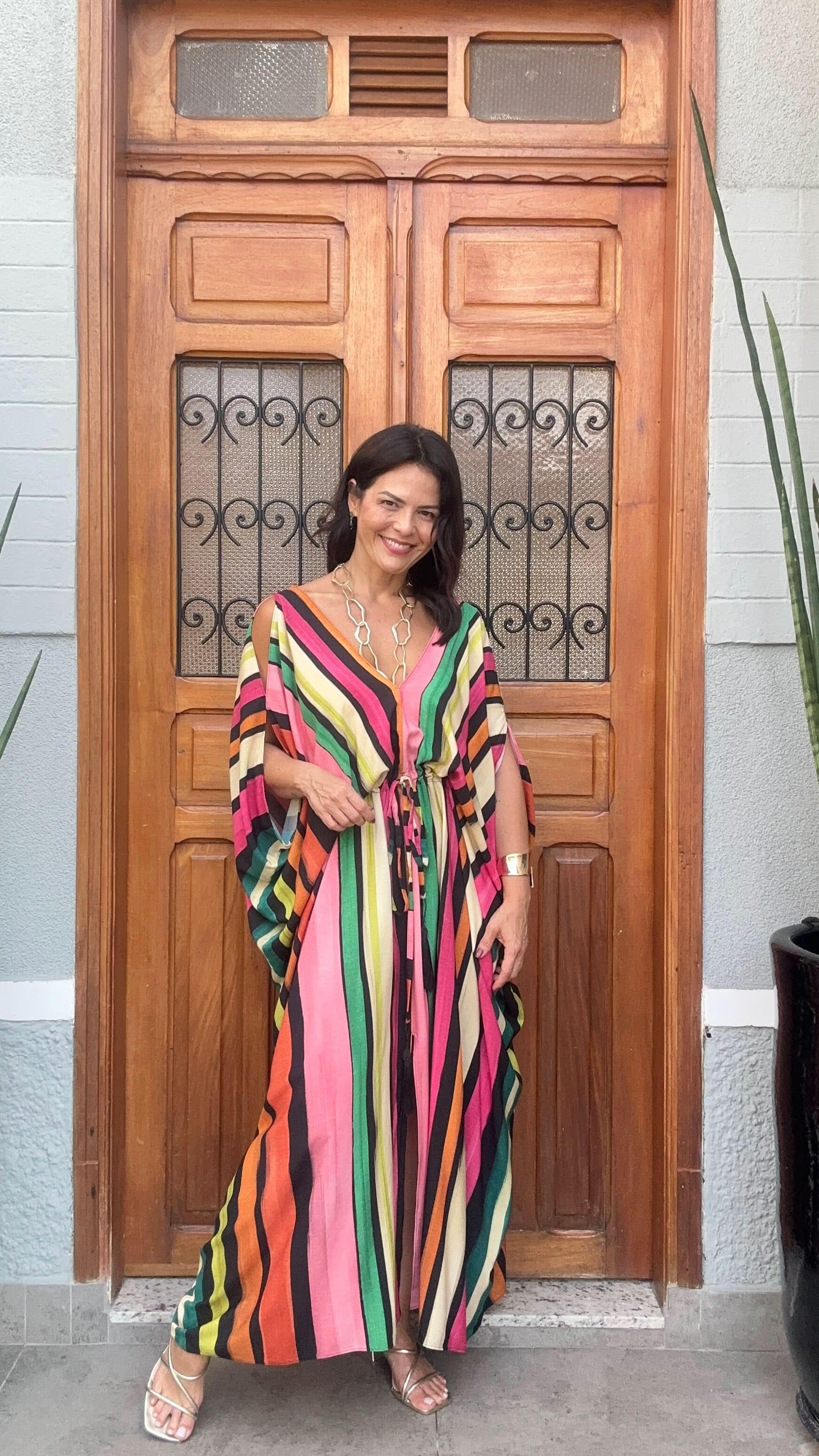 Mulher sorrindo usa kaftan longo listrado colorido em frente a porta de madeira decorada EZILDINHA