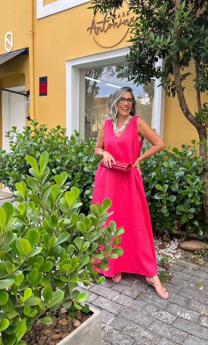 Mulher com vestido longo rosa de linho, bolsa e colar, sorrindo em jardim na frente de casa amarela EZILDINHA