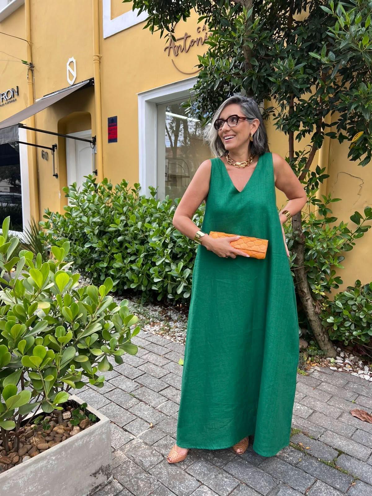Mulher sorrindo usa vestido longo verde de linho, clutch laranja, óculos e colar dourado, em ambiente urbano com plantas EZILDINHA