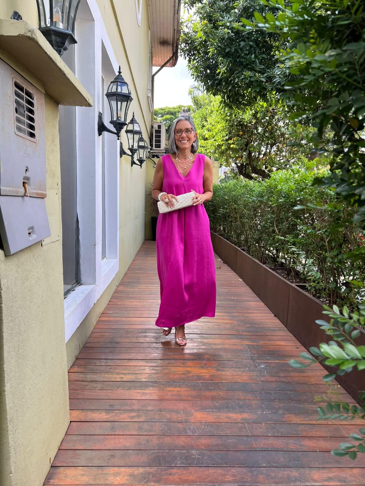 Mulher elegante com vestido longo rosa de linho, sandálias e clutch, em varanda arborizada EZILDINHA