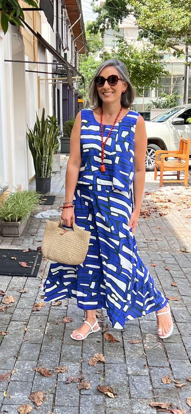 Mulher usando vestido longo estampado azul e branco, sandálias brancas e bolsa de palha, em ambiente urbano EZILDINHA