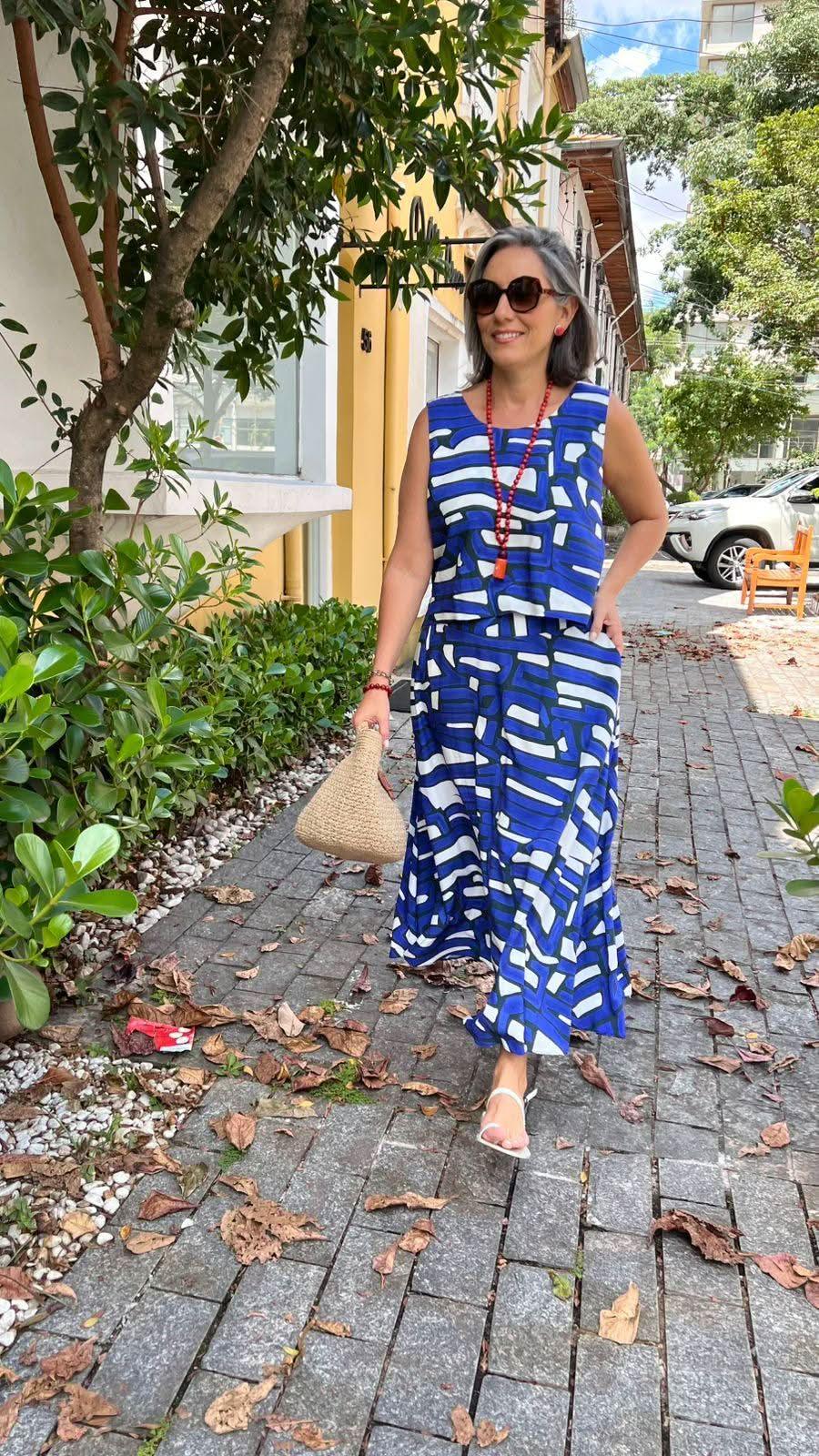 Mulher usando conjunto azul estampado, sandálias brancas e bolsa de palha, caminhando em calçada arborizada EZILDINHA