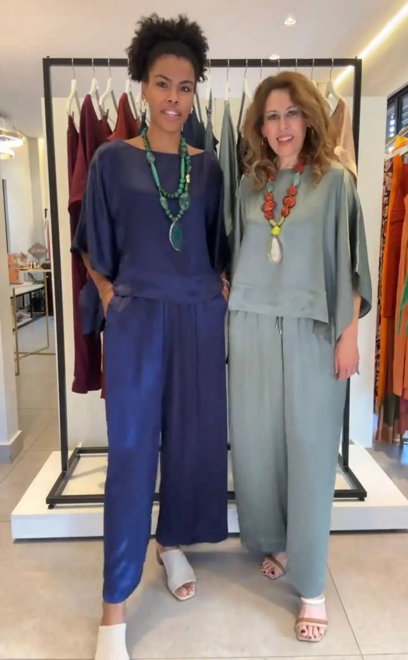 Duas mulheres usando conjunto kimono viscose elegante, uma azul marinho e outra verde, com colares grandes, em loja de roupas EZILDINHA