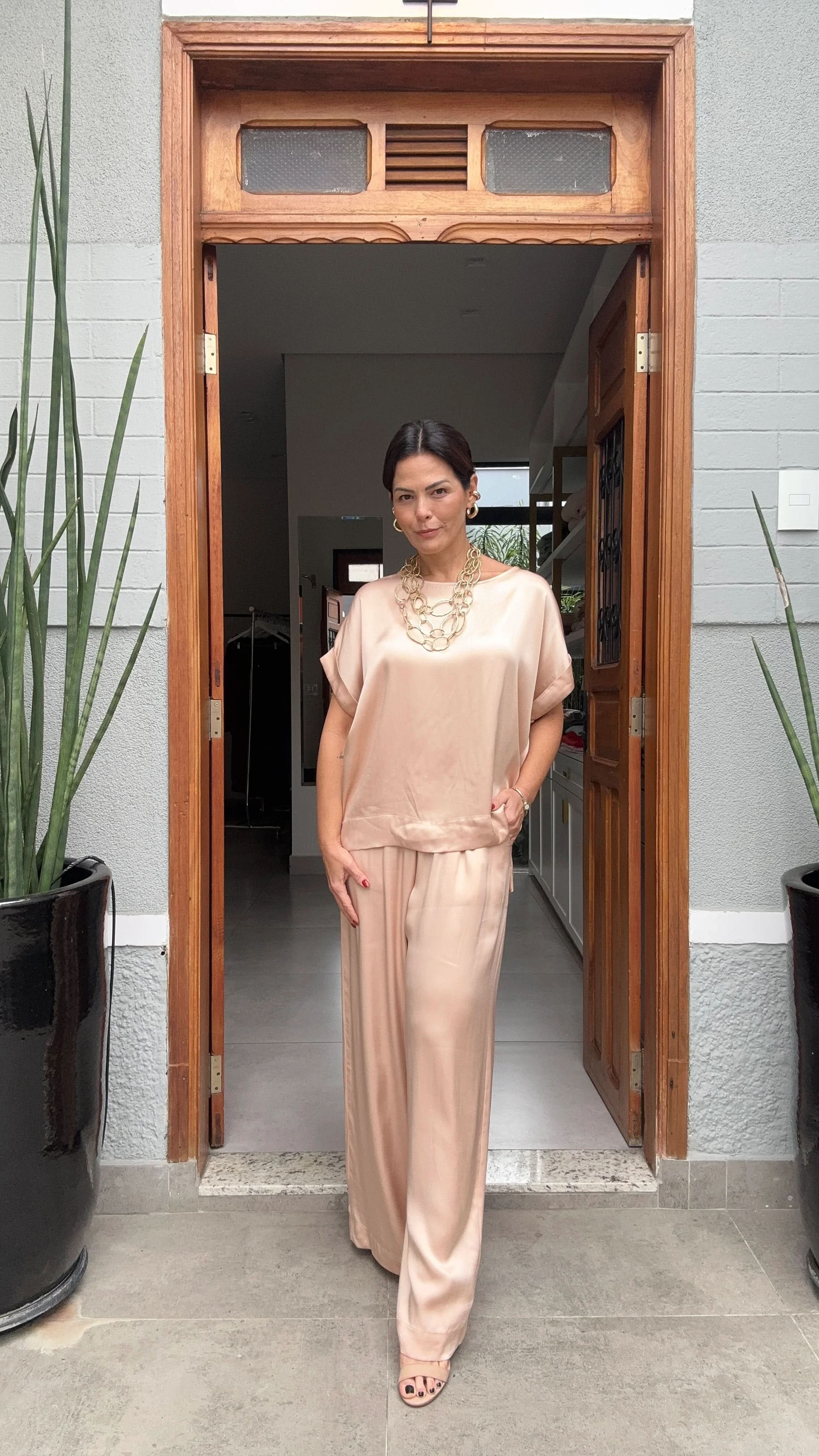Mulher usando conjunto acetinado nude de manguinha japonesa, calça ampla, colar dourado, em porta de entrada elegante EZILDINHA
