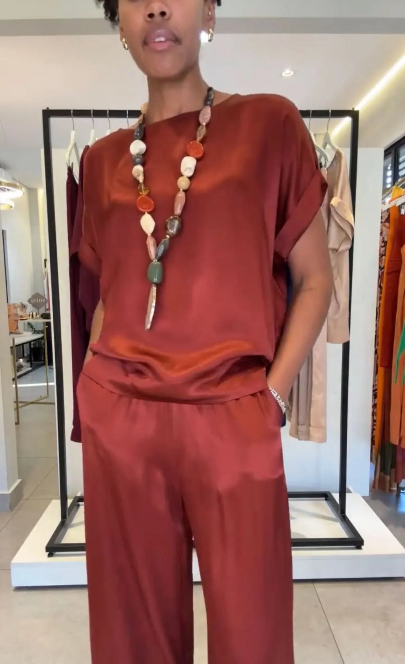 Mulher veste conjunto pantalona em tecido acetinado vinho, com manga japonesa, colar de pedras coloridas, em loja de moda feminina EZILDINHA