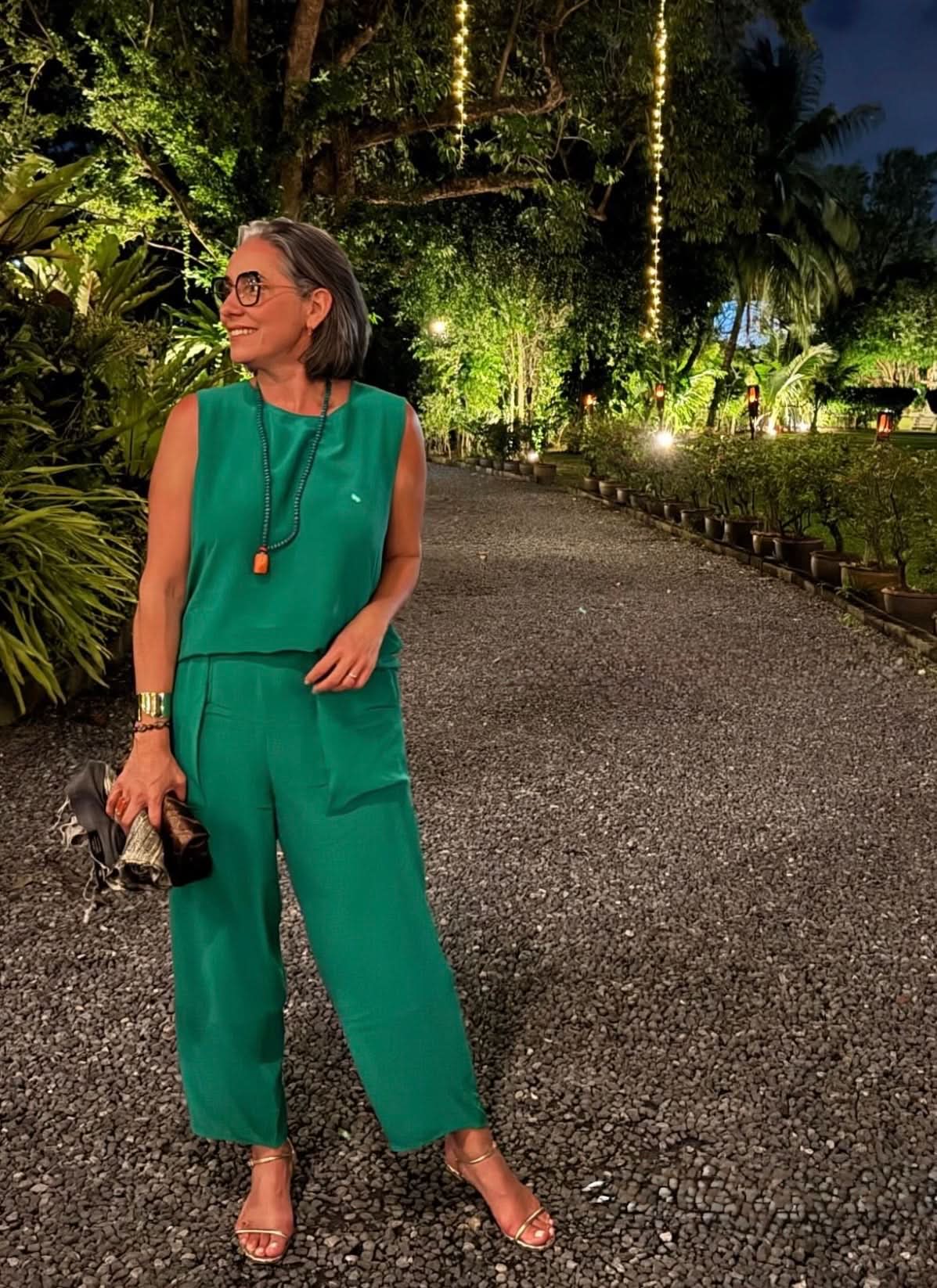 Mulher usando conjunto verde de seda, calça e blusa sem mangas, em um jardim iluminado à noite EZILDINHA