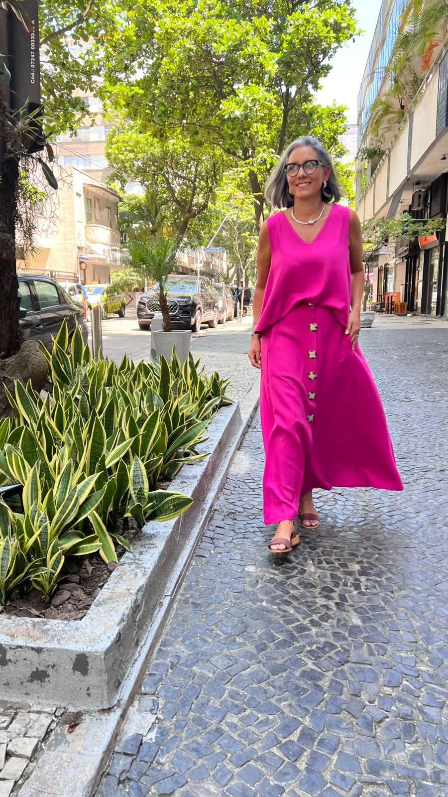 Mulher com cabelo grisalho usando conjunto de saia e regata rosa vivo, andando na calçada arborizada EZILDINHA