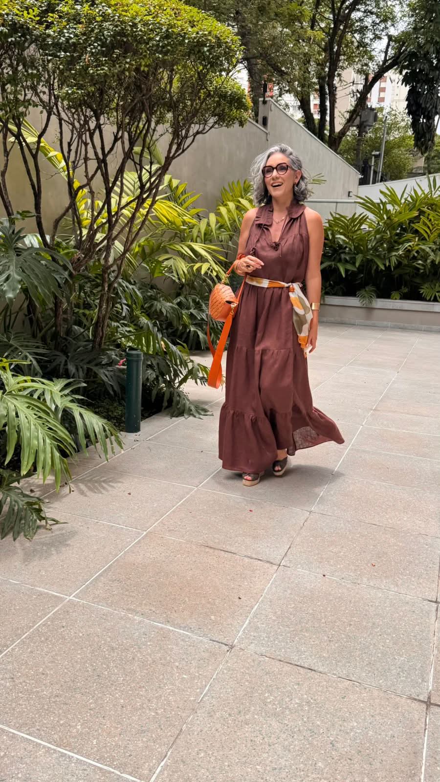 Mulher usando vestido longo de linho marrom, com lenço estampado, bolsa laranja e sandálias, caminhando em jardim urbano EZILDINHA