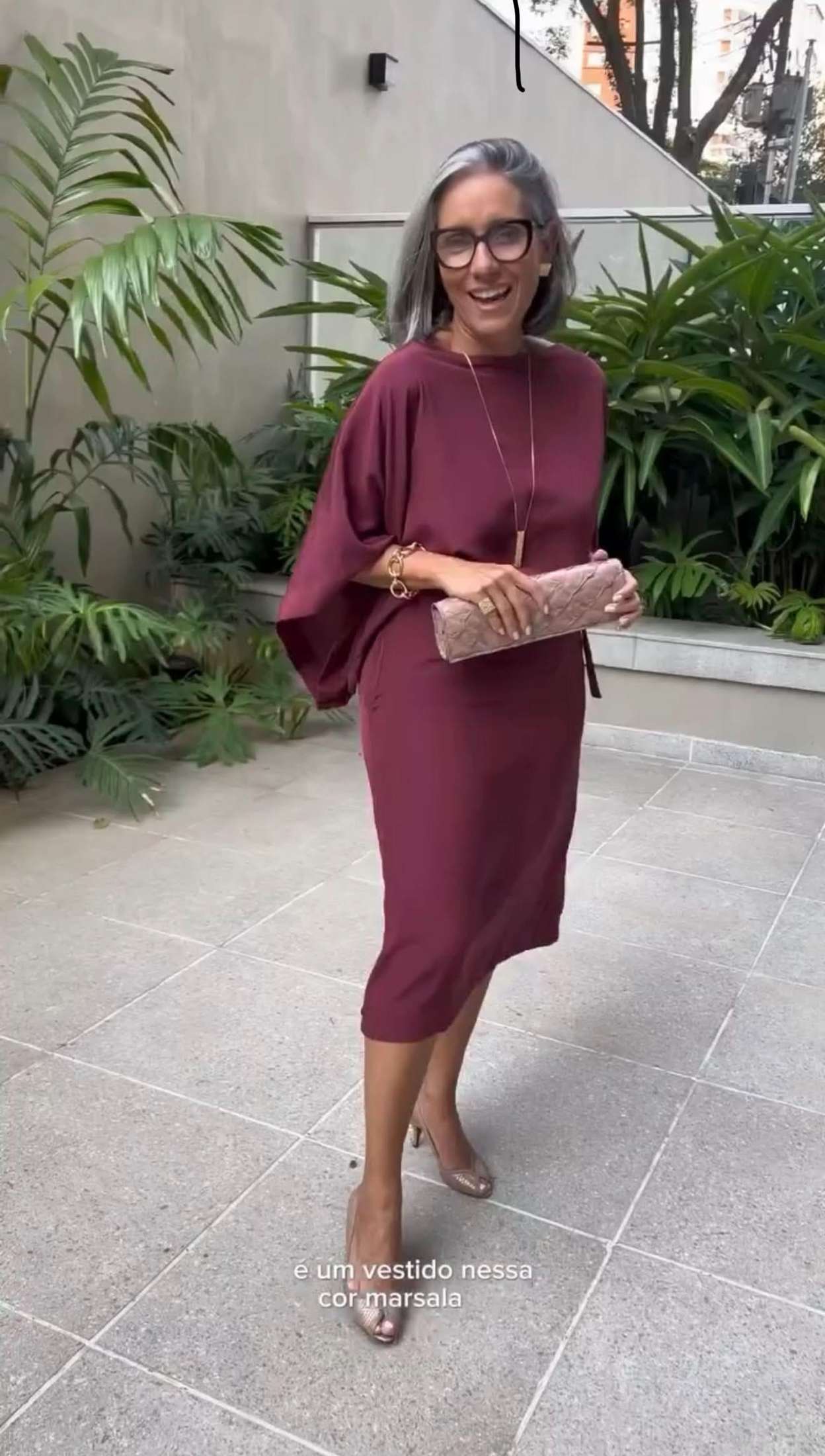 Mulher usando vestido midi marsala de manga ampla e salto, segurando clutch bege em área externa com plantas EZILDINHA