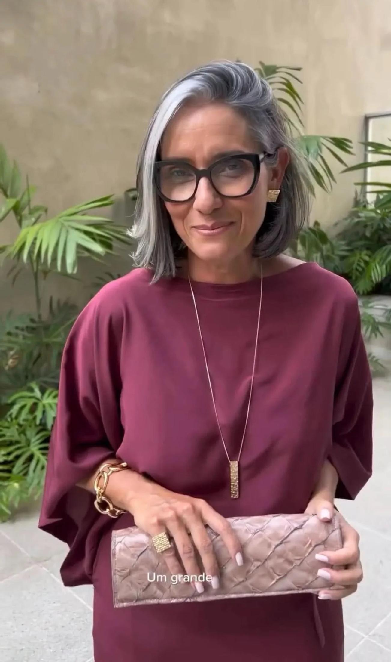 Mulher madura com cabelo grisalho usando vestido vinho e acessórios dourados, segurando clutch bege EZILDINHA