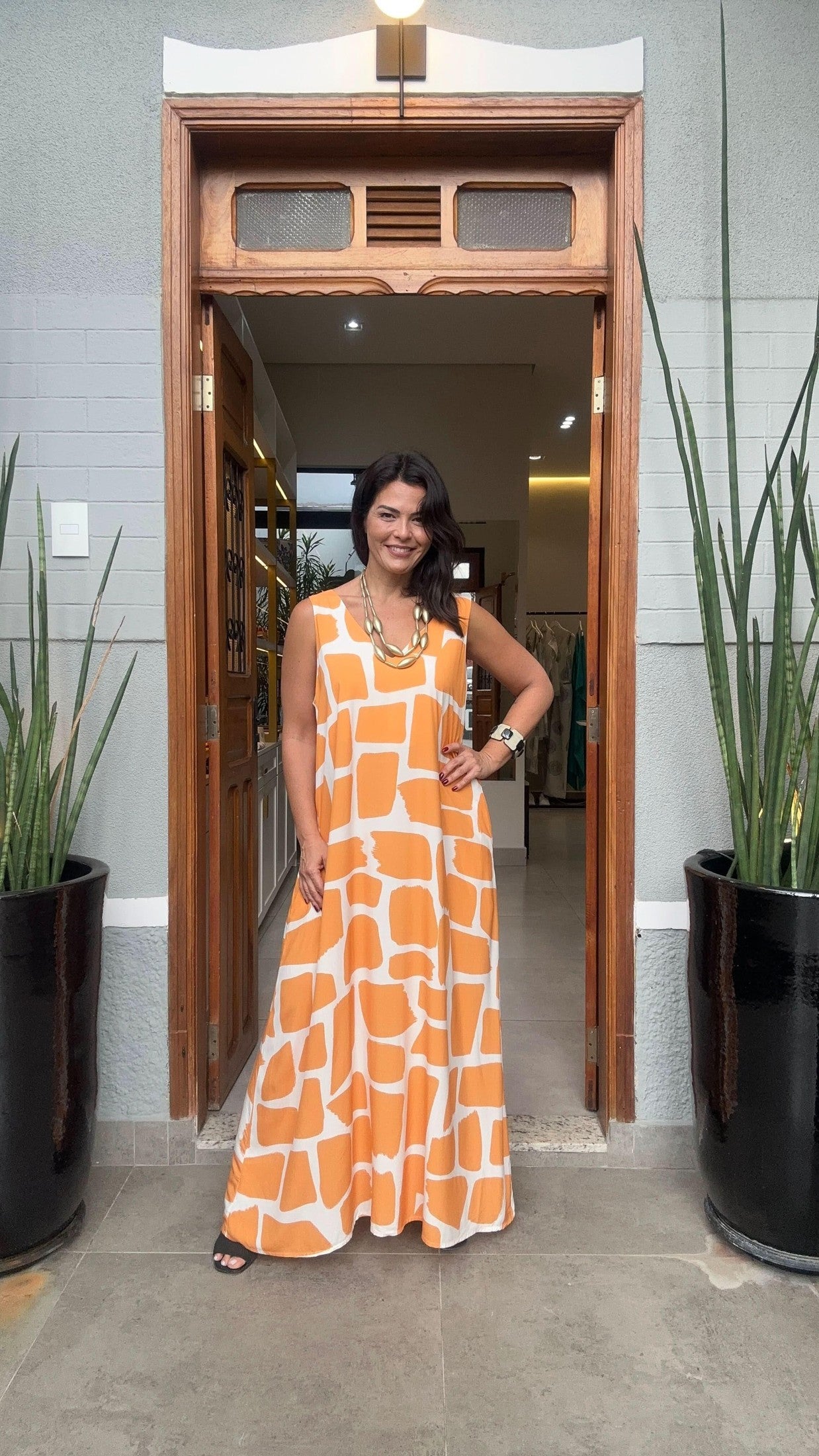 Mulher usando vestido longo estampado laranja e branco, em porta de entrada com plantas ao lado EZILDINHA