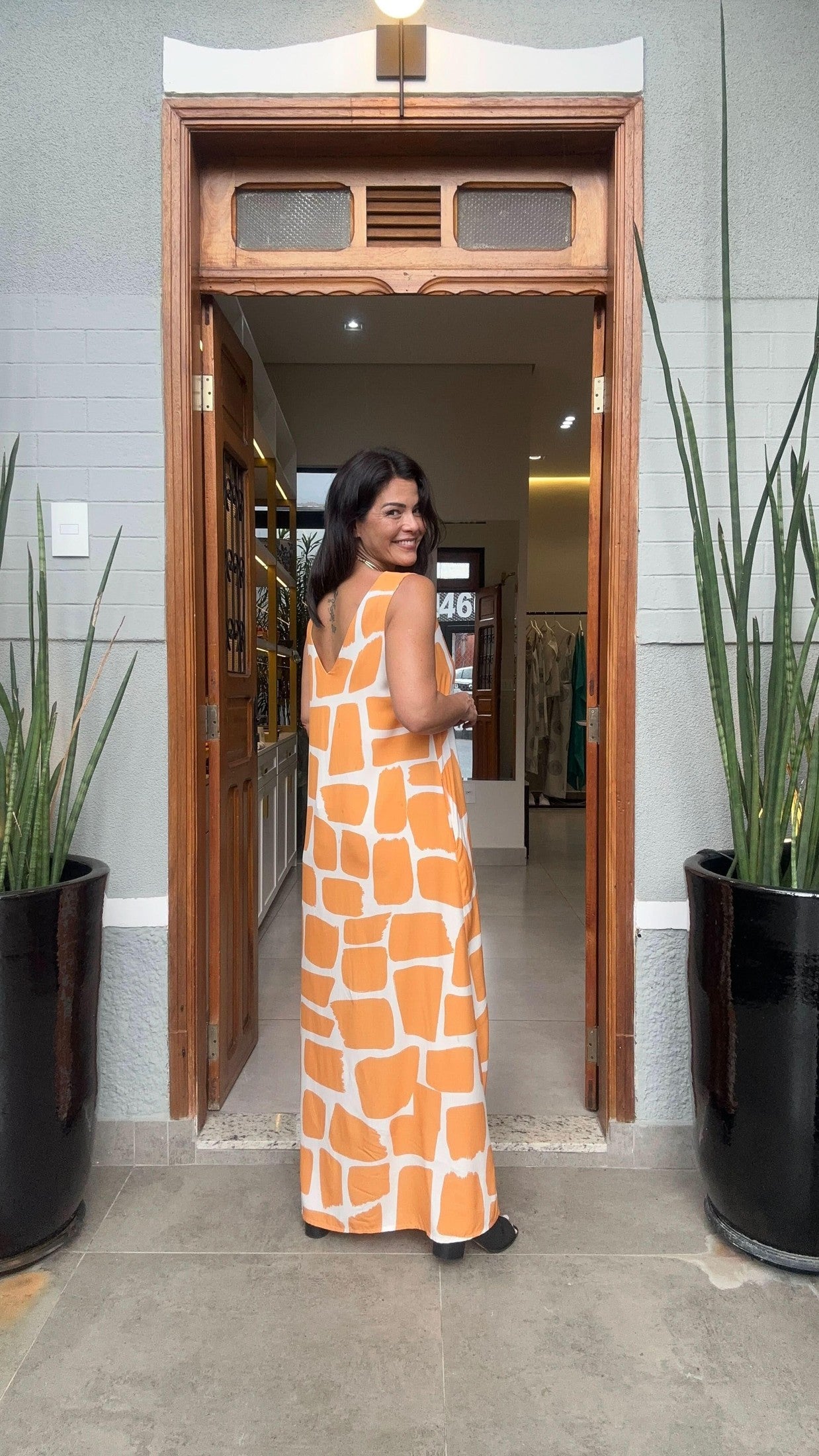 Mulher usando vestido longo estampado laranja e branco, em frente a uma porta com vasos de plantas EZILDINHA