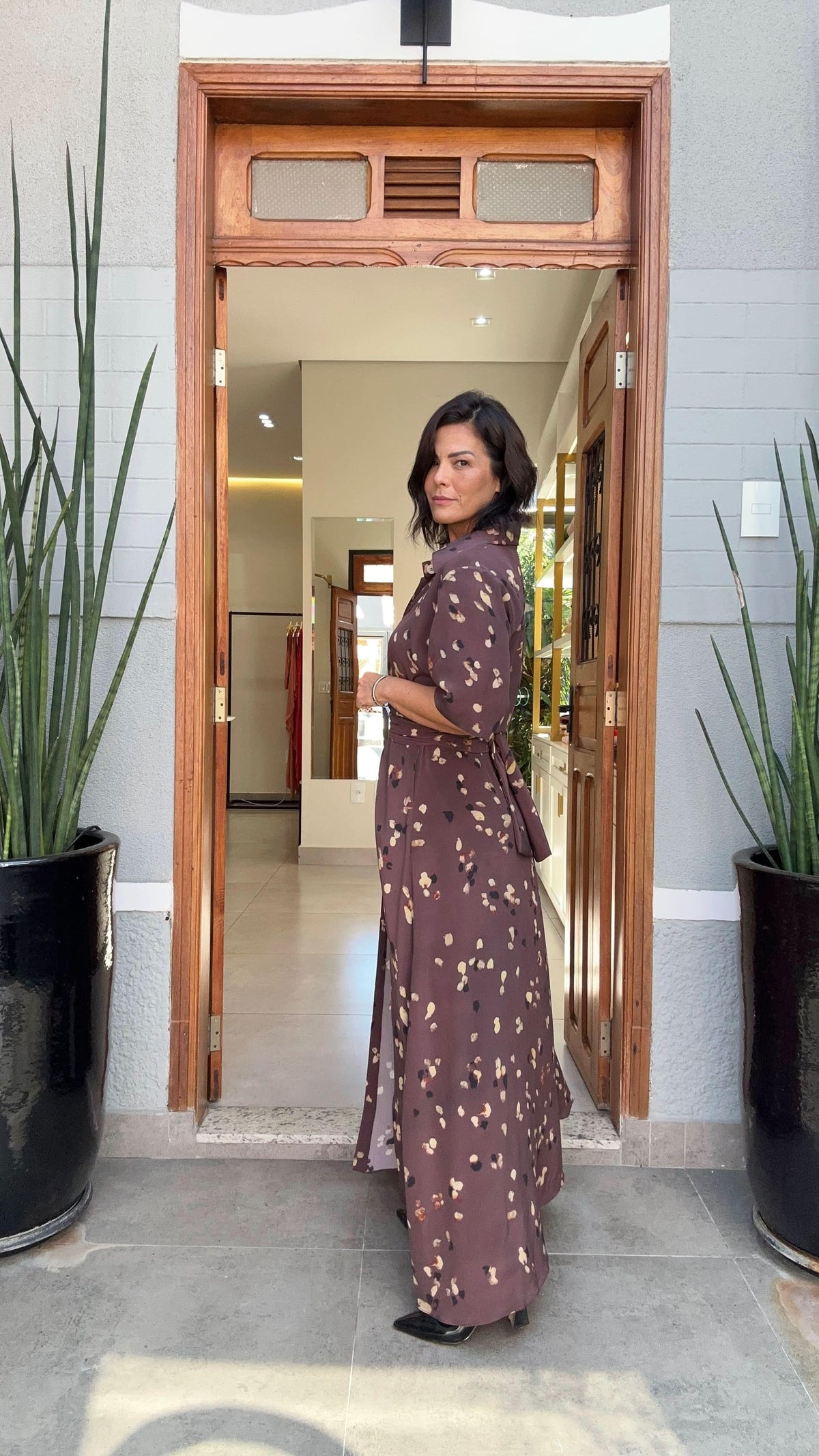 Mulher usando vestido longo estampado de seda marrom, em frente a porta de madeira, entre plantas decorativas EZILDINHA