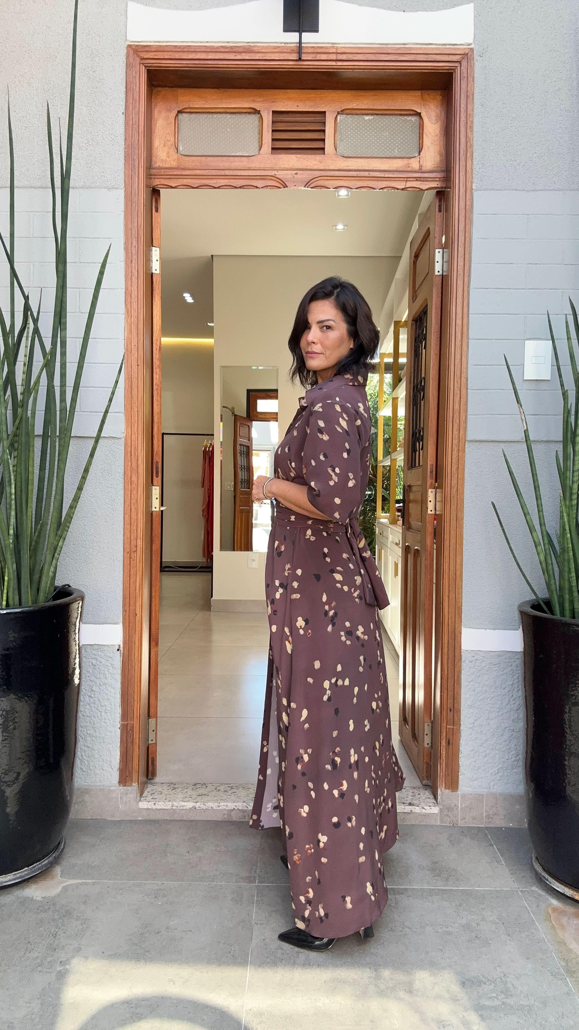 Mulher usando vestido longo estampado de seda marrom, em frente a porta de madeira, entre plantas decorativas EZILDINHA