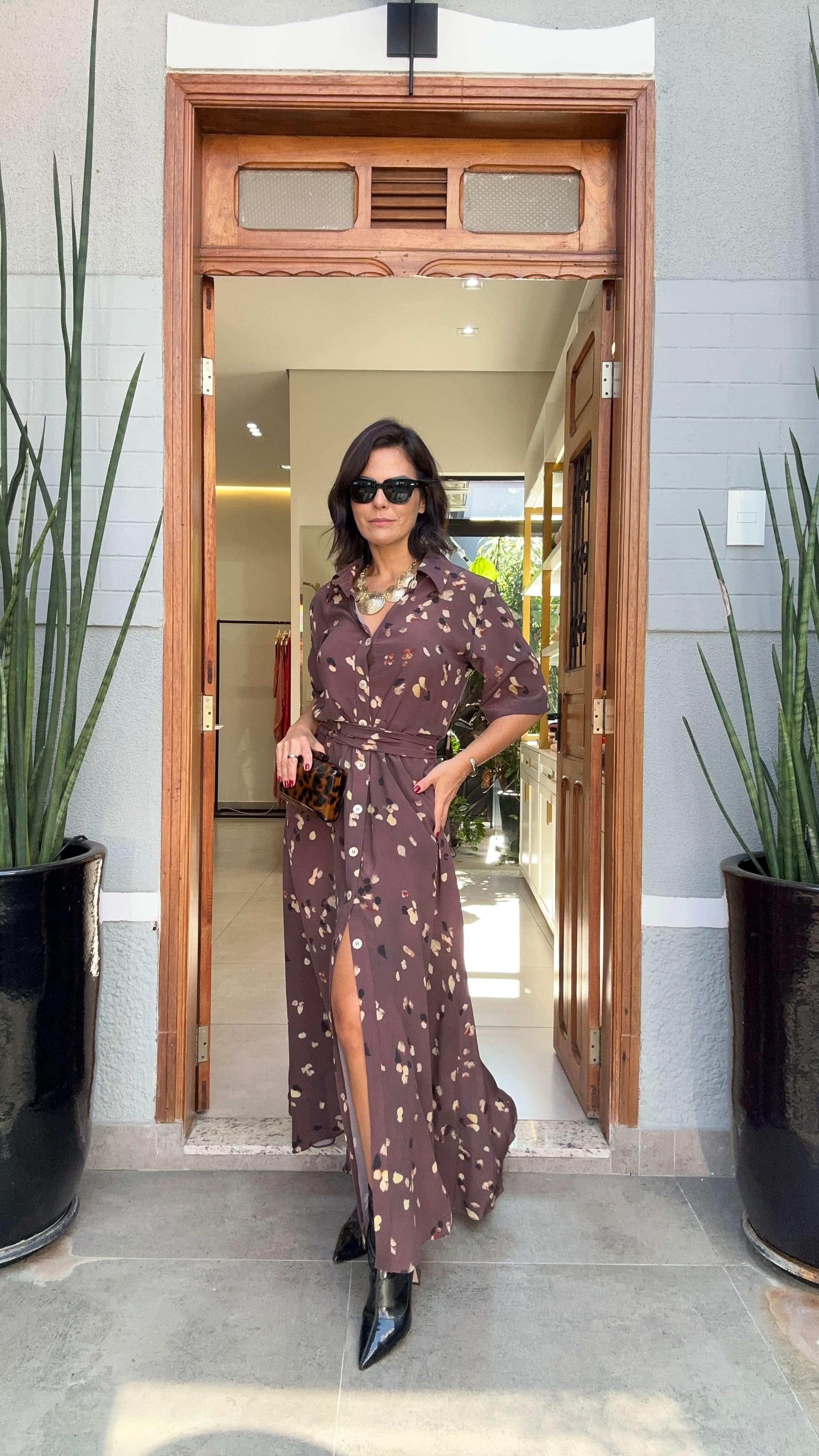 Mulher usando vestido longo estampado marrom, óculos escuros e botas pretas, em entrada de casa EZILDINHA