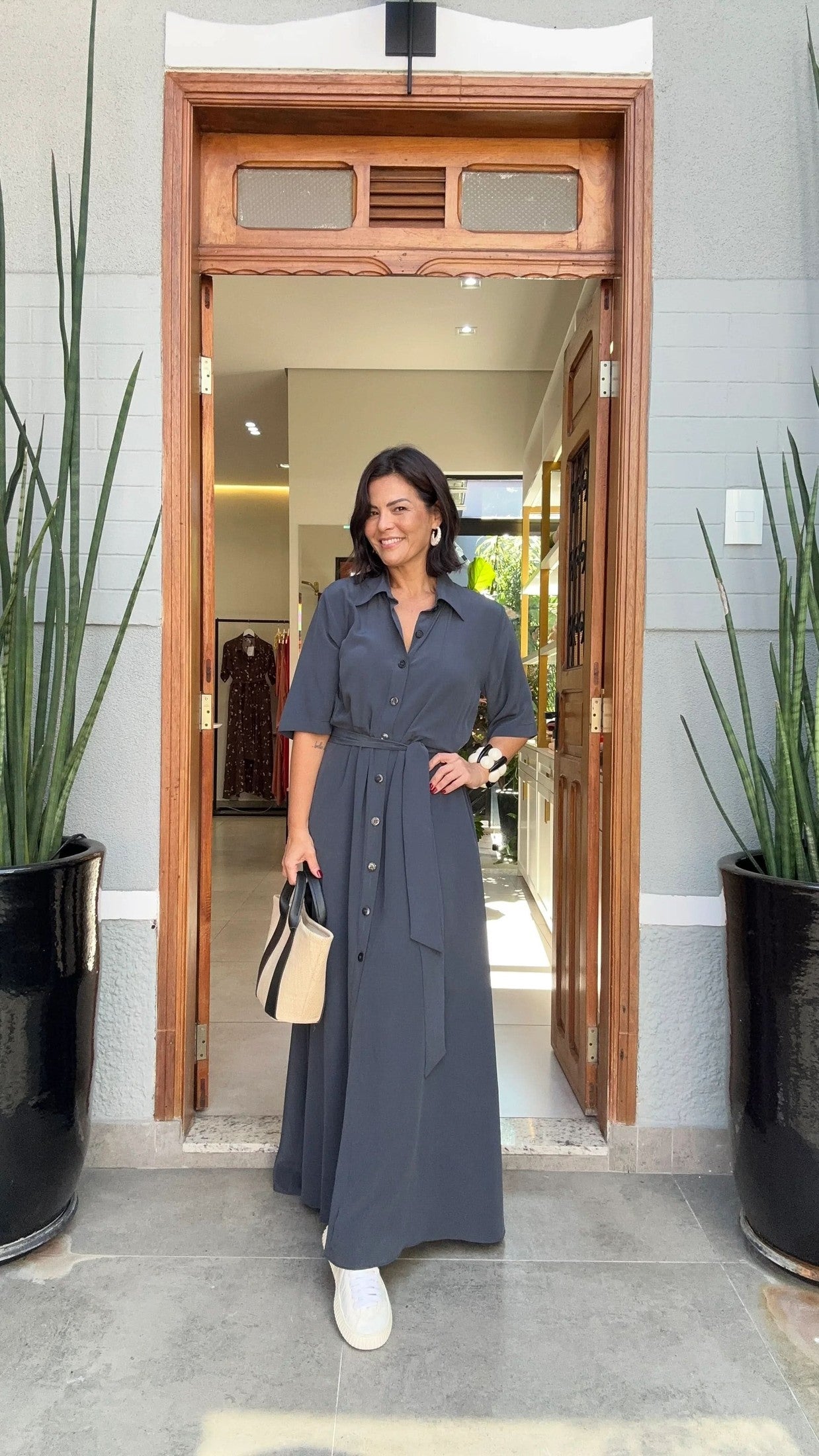 Mulher usando vestido longo azul-marinho de linho, segurando bolsa bege, em entrada de loja EZILDINHA