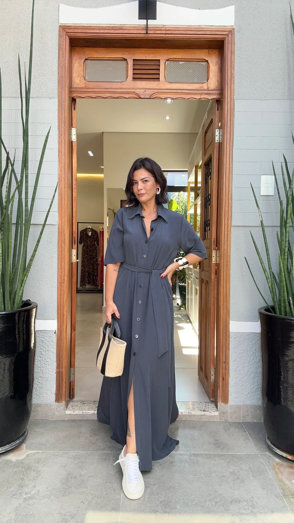 Mulher usando vestido longo azul de botões, bolsa de palha e tênis branco na entrada de loja EZILDINHA