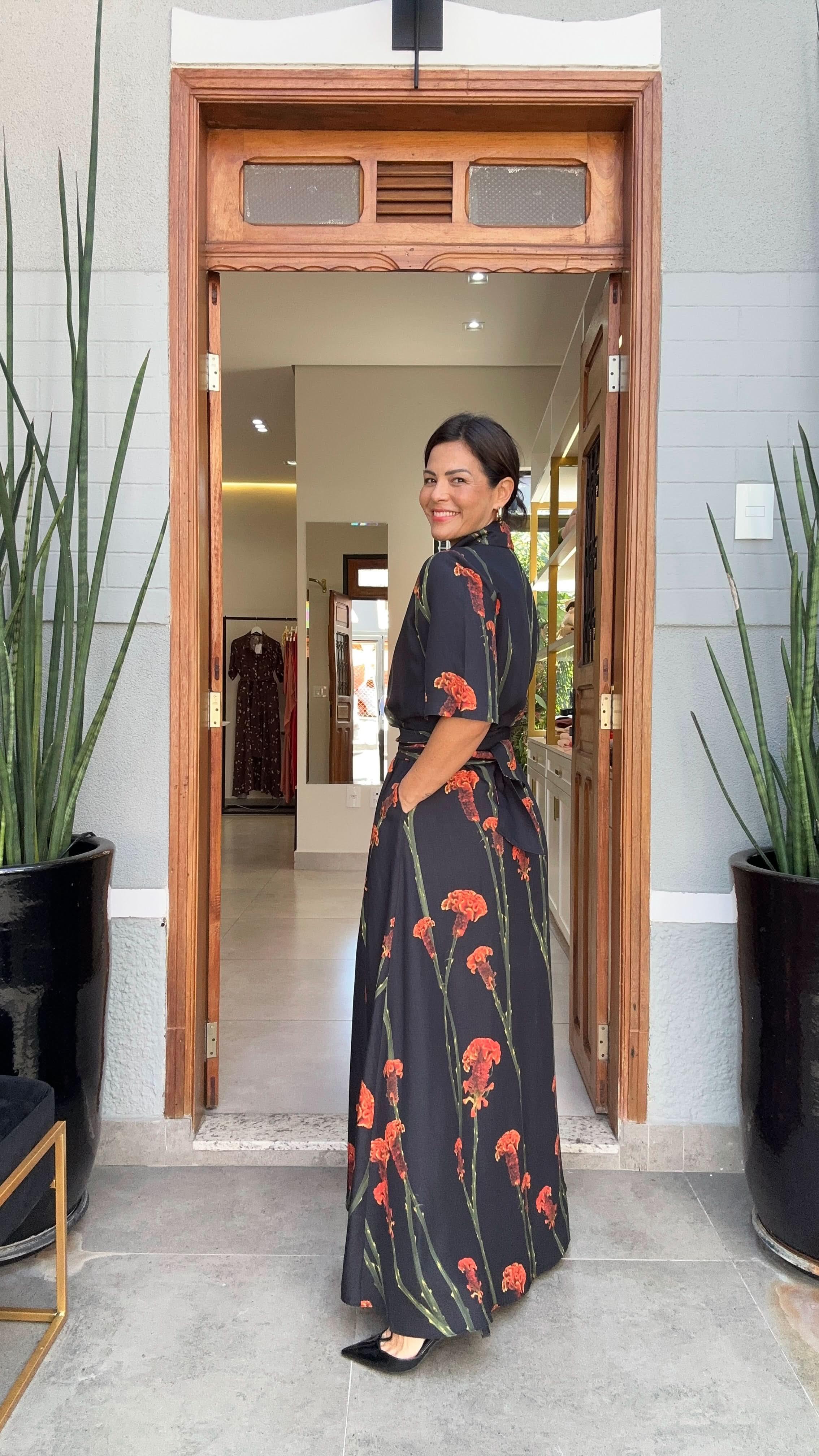 Mulher sorrindo usando vestido longo preto com estampa floral vermelha, de linho, em frente a uma porta EZILDINHA