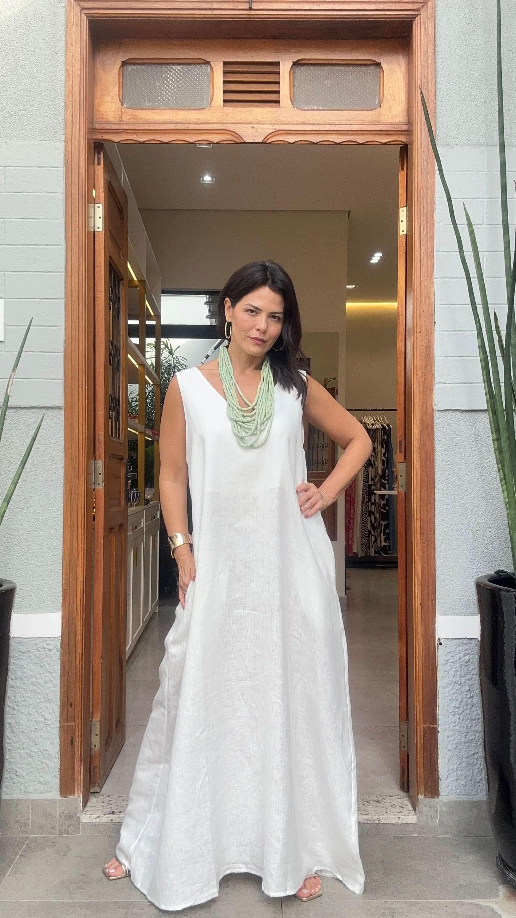 Mulher usando vestido longo branco de linho, colar verde, em entrada elegante de loja EZILDINHA