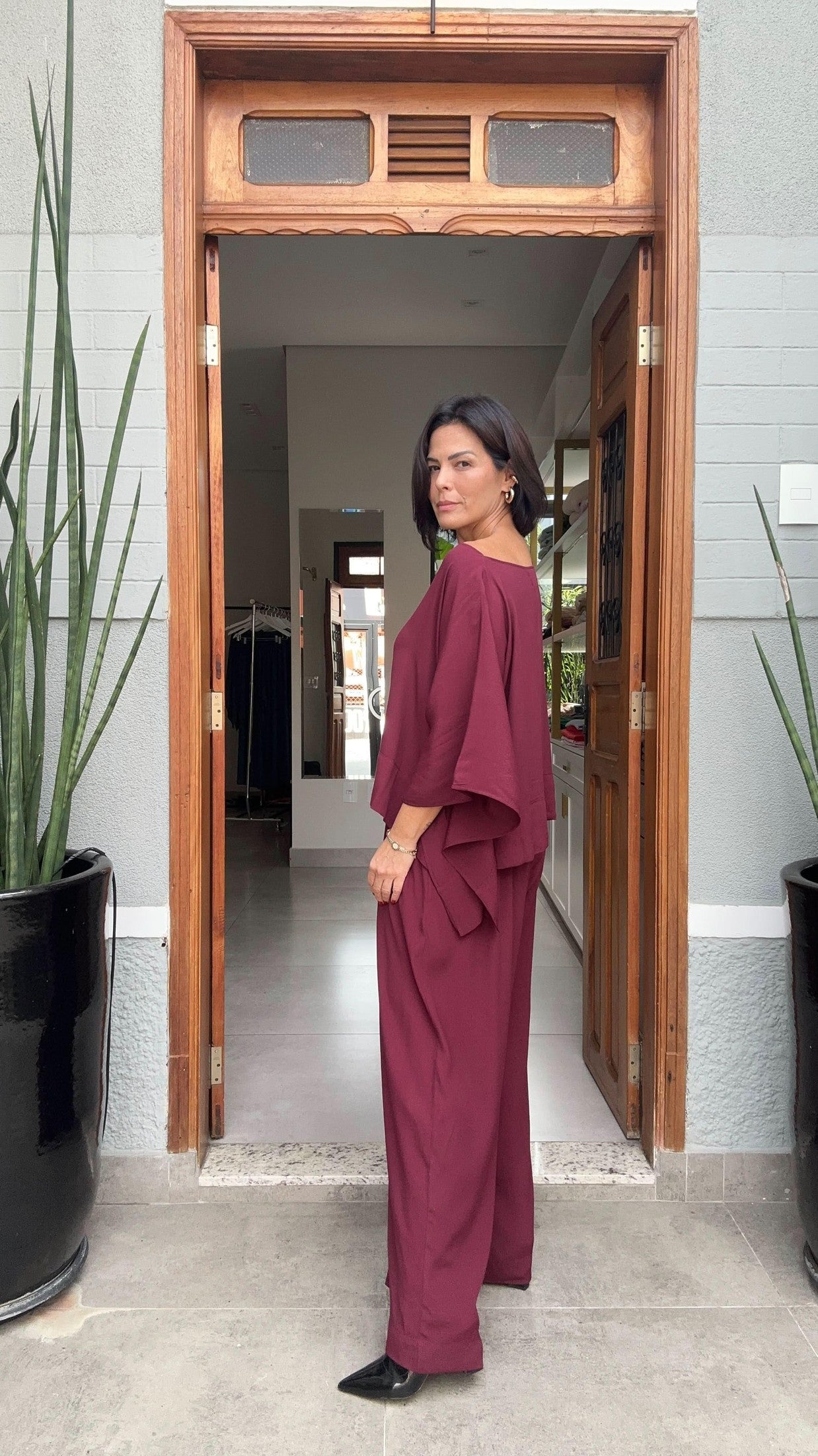 Mulher usando conjunto de linho vinho, em pé na porta de entrada moderna, plantas ao fundo EZILDINHA