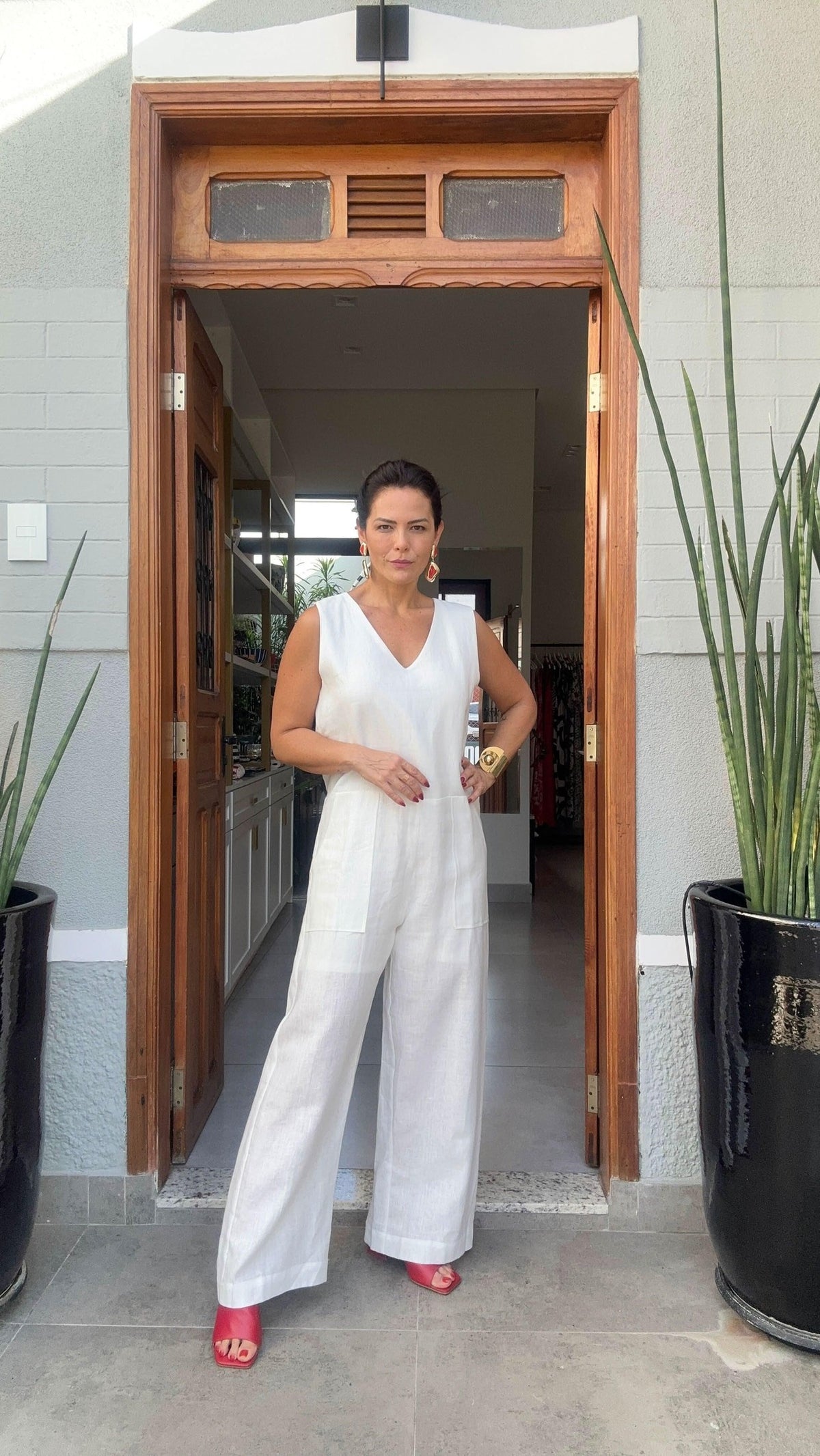Mulher usando macacão branco de linho, sandálias vermelhas, em entrada moderna com plantas EZILDINHA