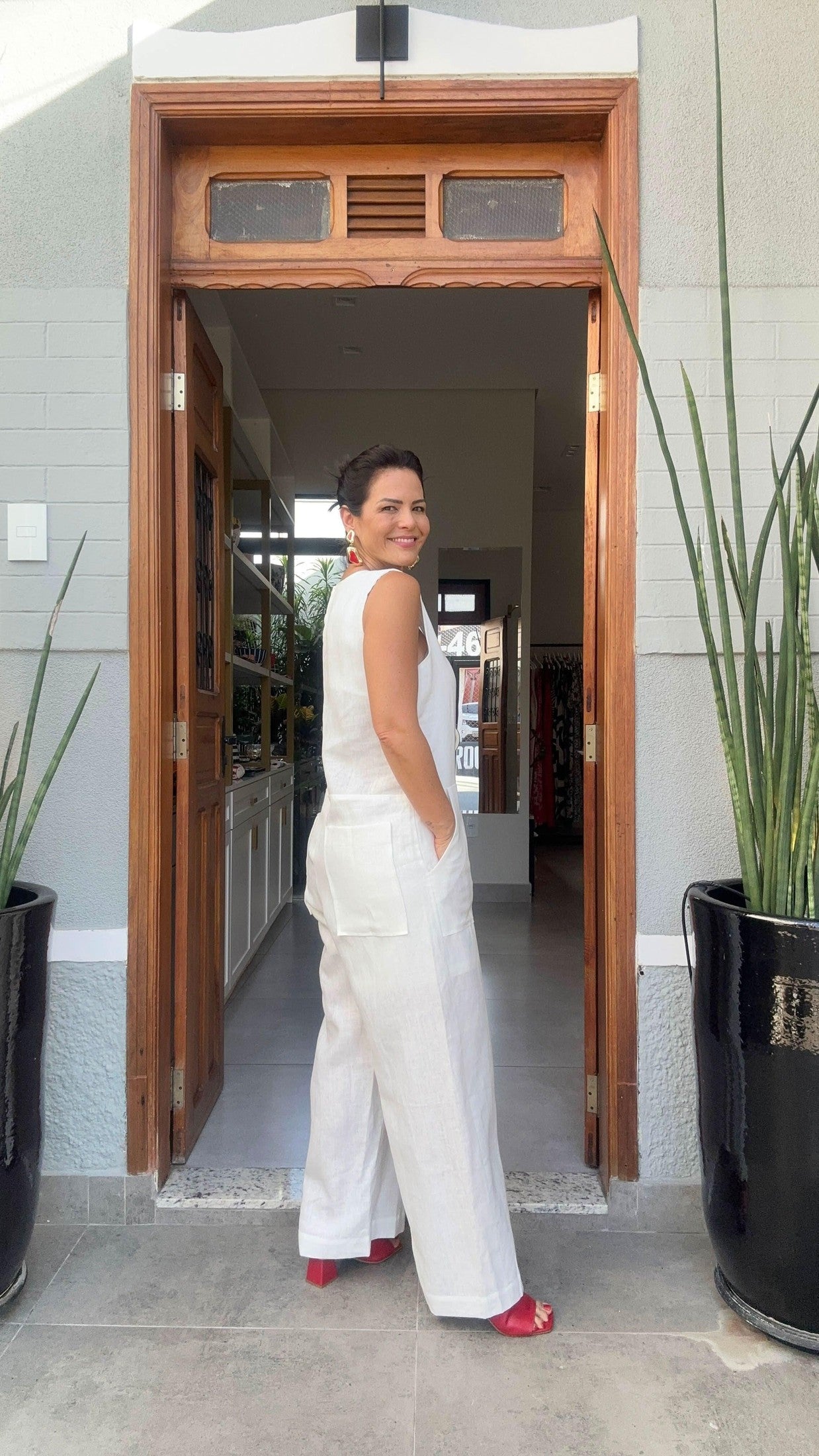 Mulher sorrindo usando macacão branco de linho e sandálias vermelhas na entrada de loja elegante EZILDINHA