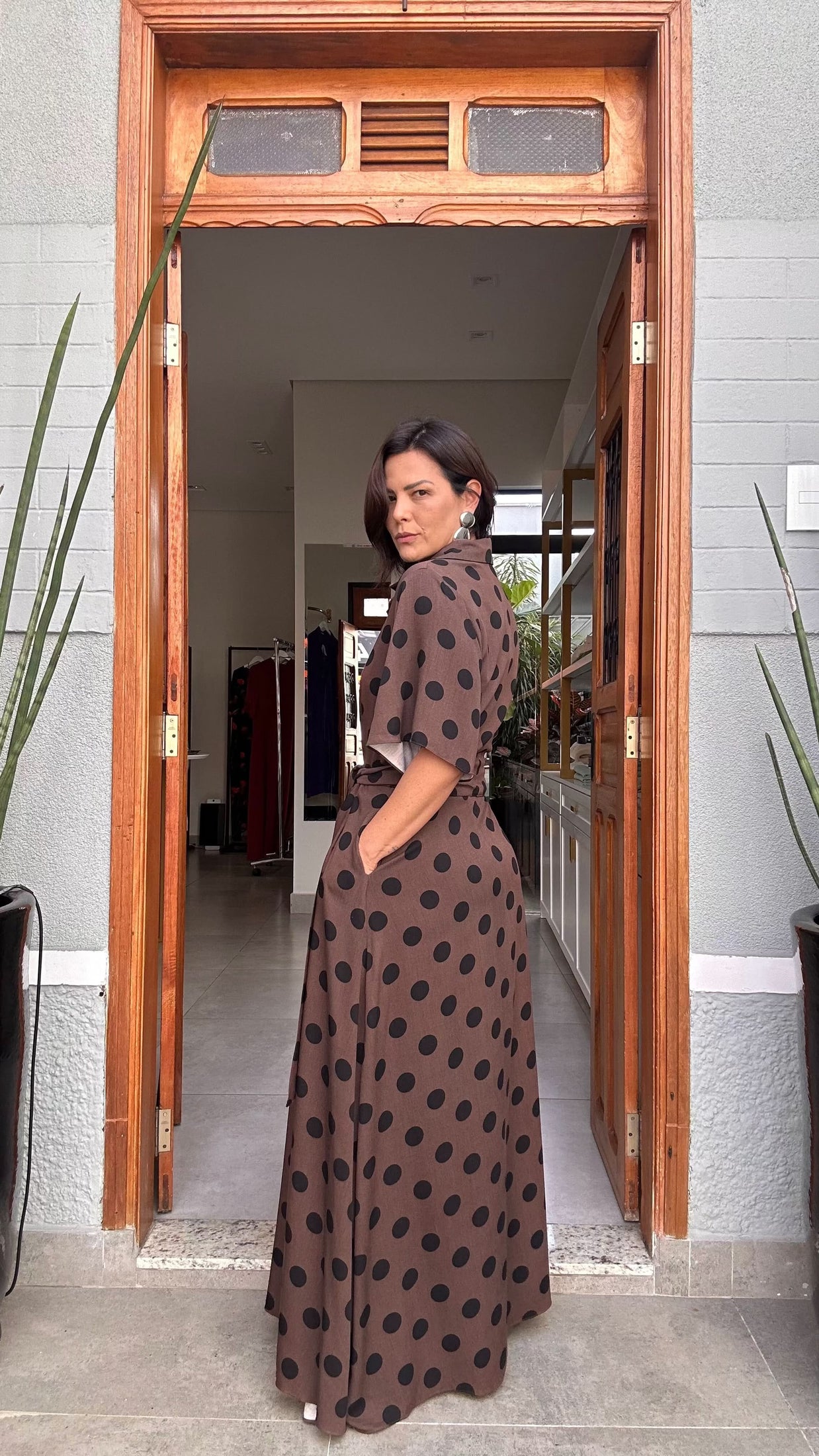 Mulher usando vestido longo marrom com estampa de poá preto, em pé em porta de entrada EZILDINHA