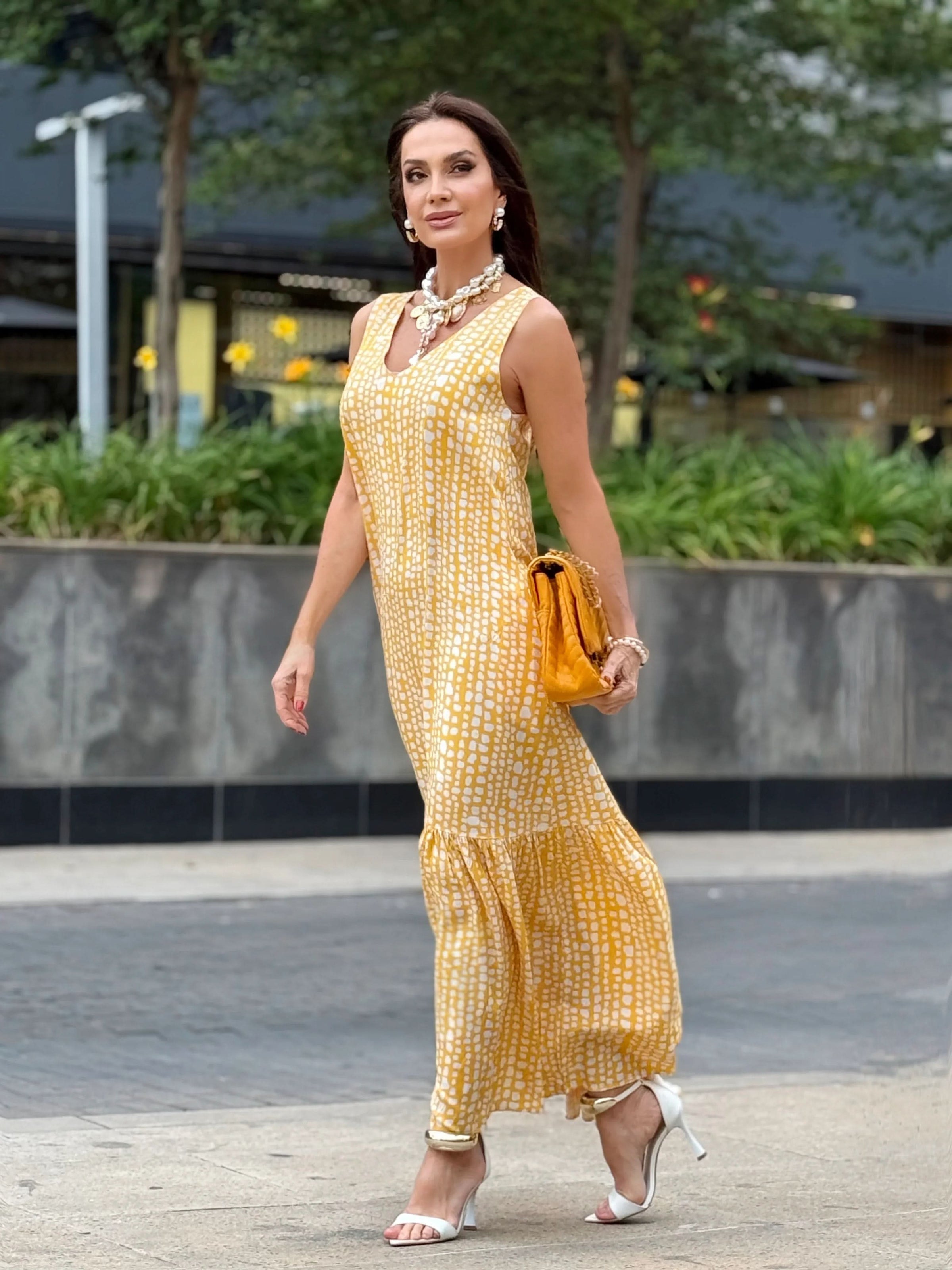Mulher usando vestido longo amarelo estampado de seda, bolsa mostarda e sandálias brancas, ao ar livre EZILDINHA