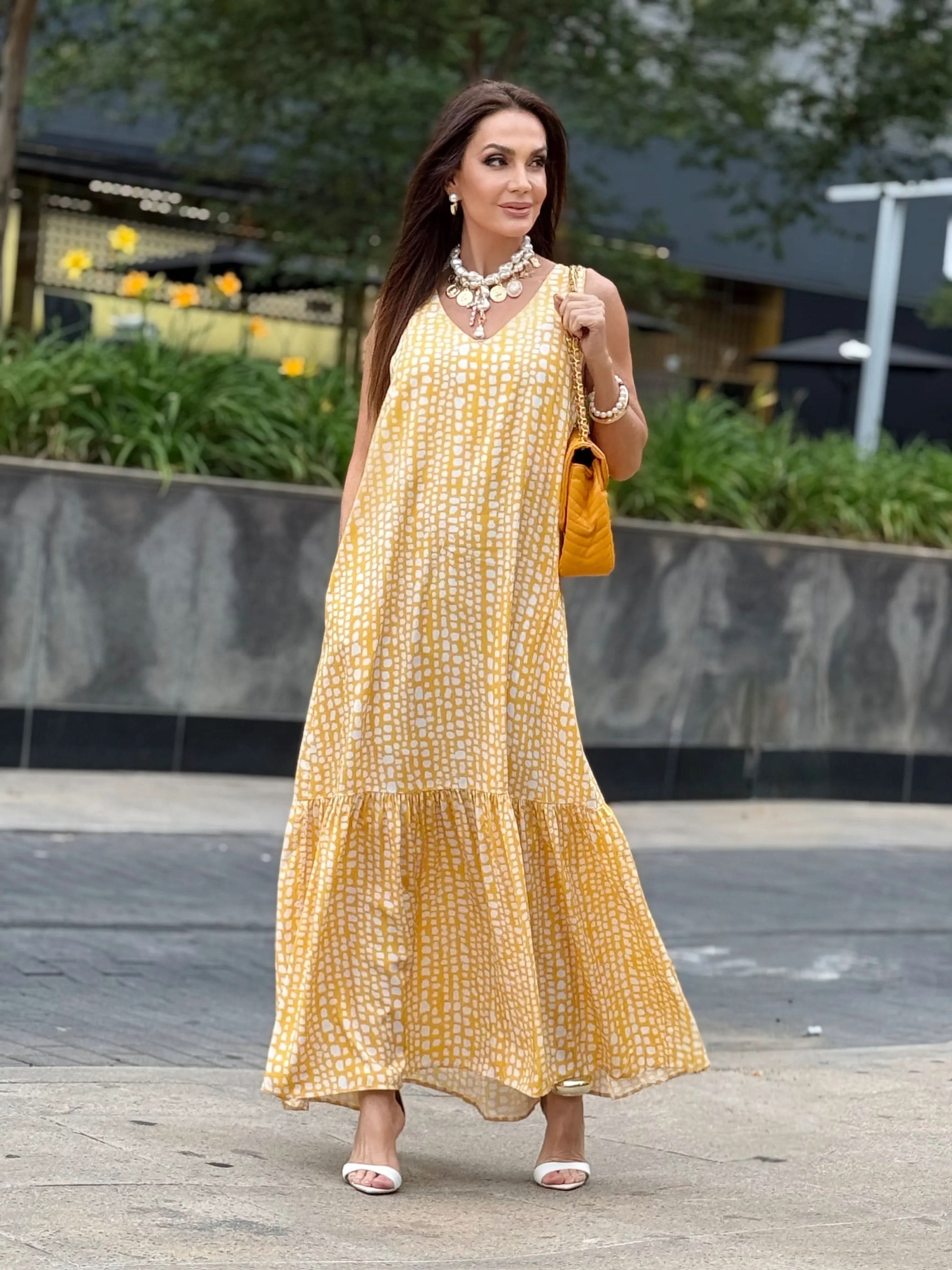 Mulher com vestido longo amarelo estampado, bolsa amarela e sandália branca ao ar livre EZILDINHA