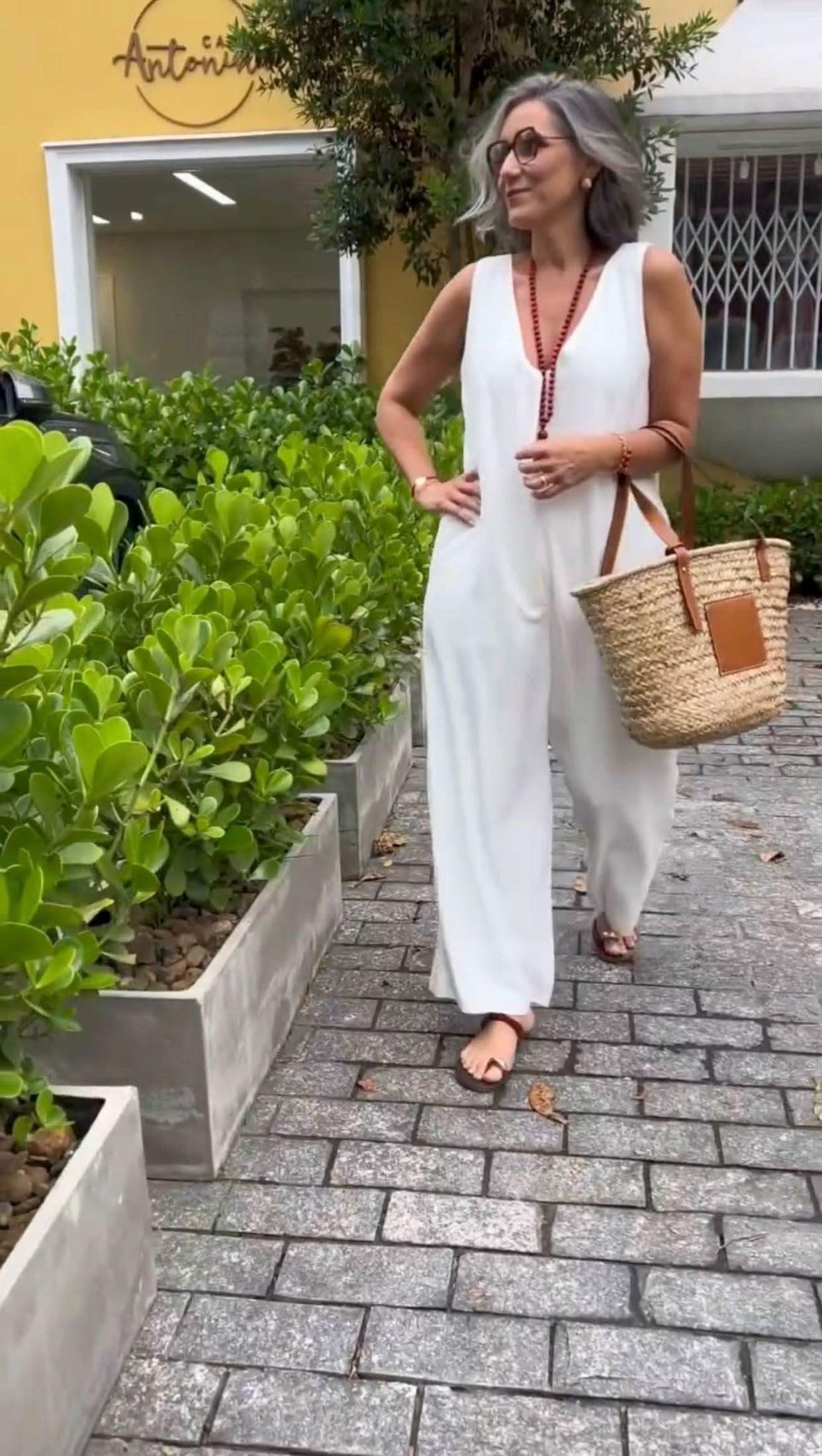 Mulher madura usando macacão branco de linho, sandálias, bolsa de palha e colar, caminhando ao ar livre EZILDINHA