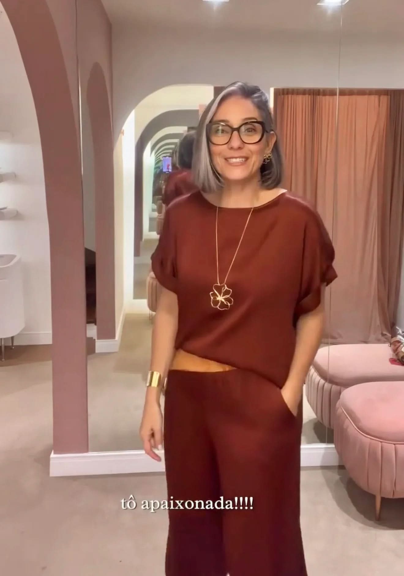 Mulher sorrindo veste conjunto de linho vinho, acessórios dourados, em loja elegante EZILDINHA