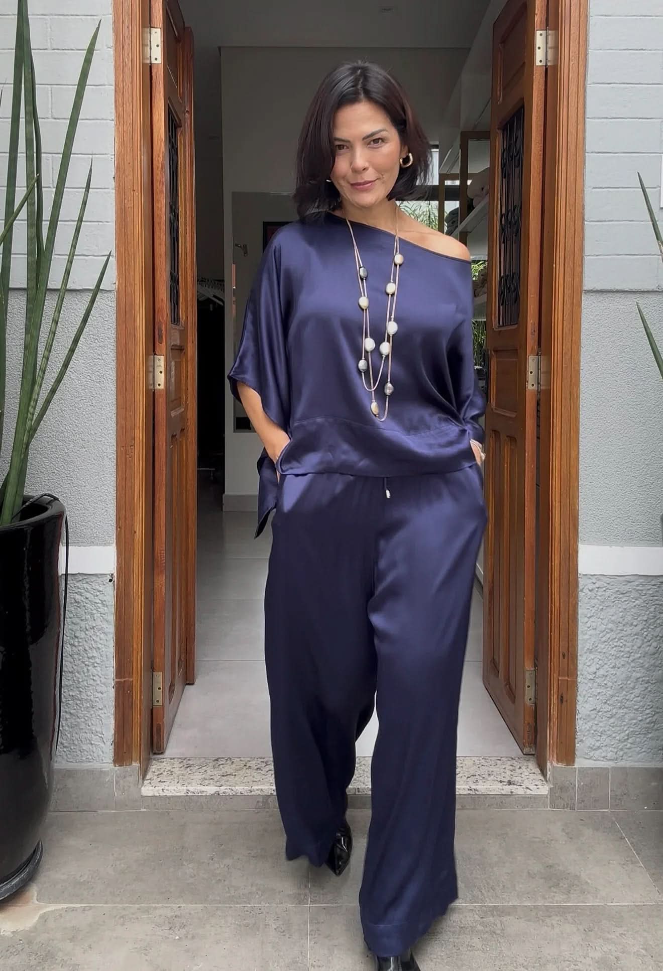 Mulher vestindo conjunto de seda azul marinho, com colar longo, em ambiente moderno EZILDINHA