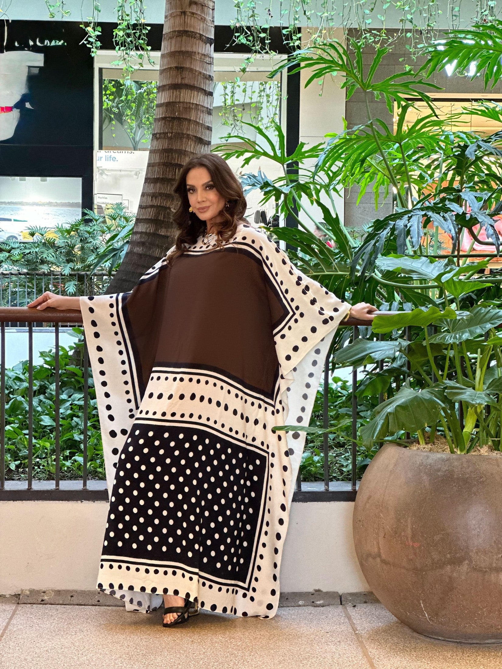 Mulher usando kaftan longo de viscose marrom com estampa de poá em ambiente com plantas tropicais EZILDINHA