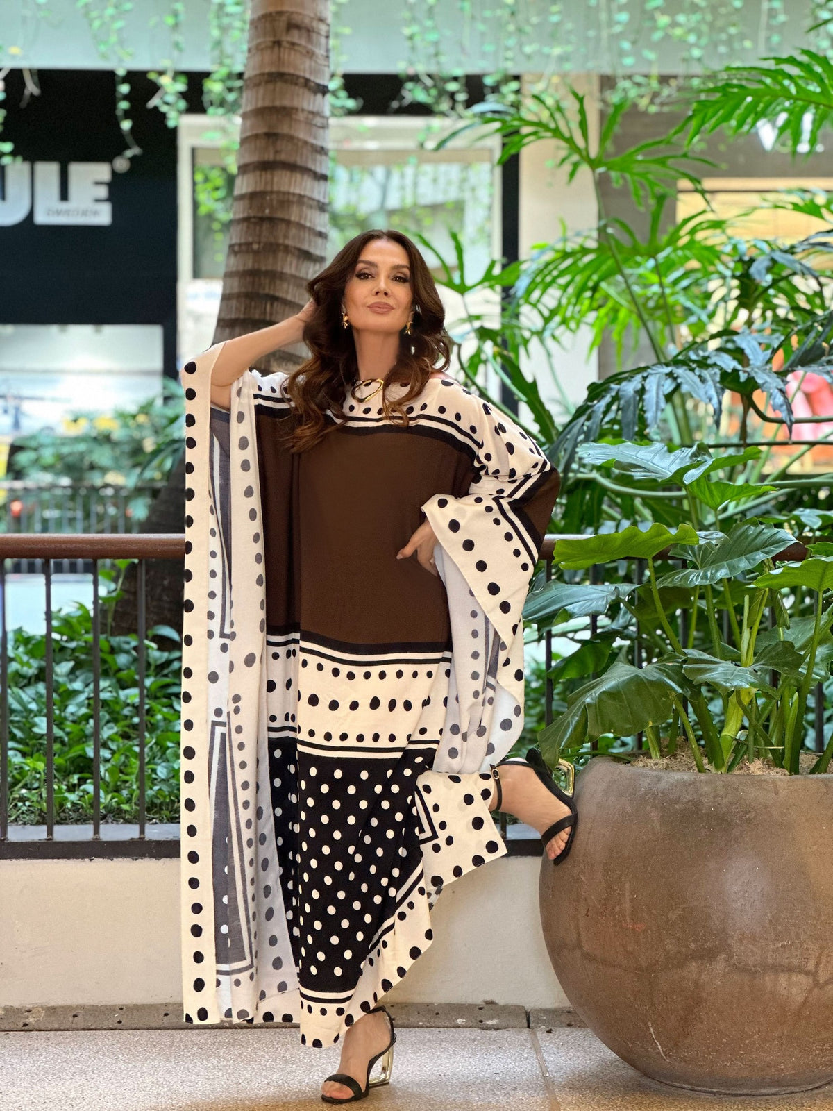 Mulher usando kaftan longo de viscose marrom e branco com estampa de poá, em ambiente com plantas EZILDINHA