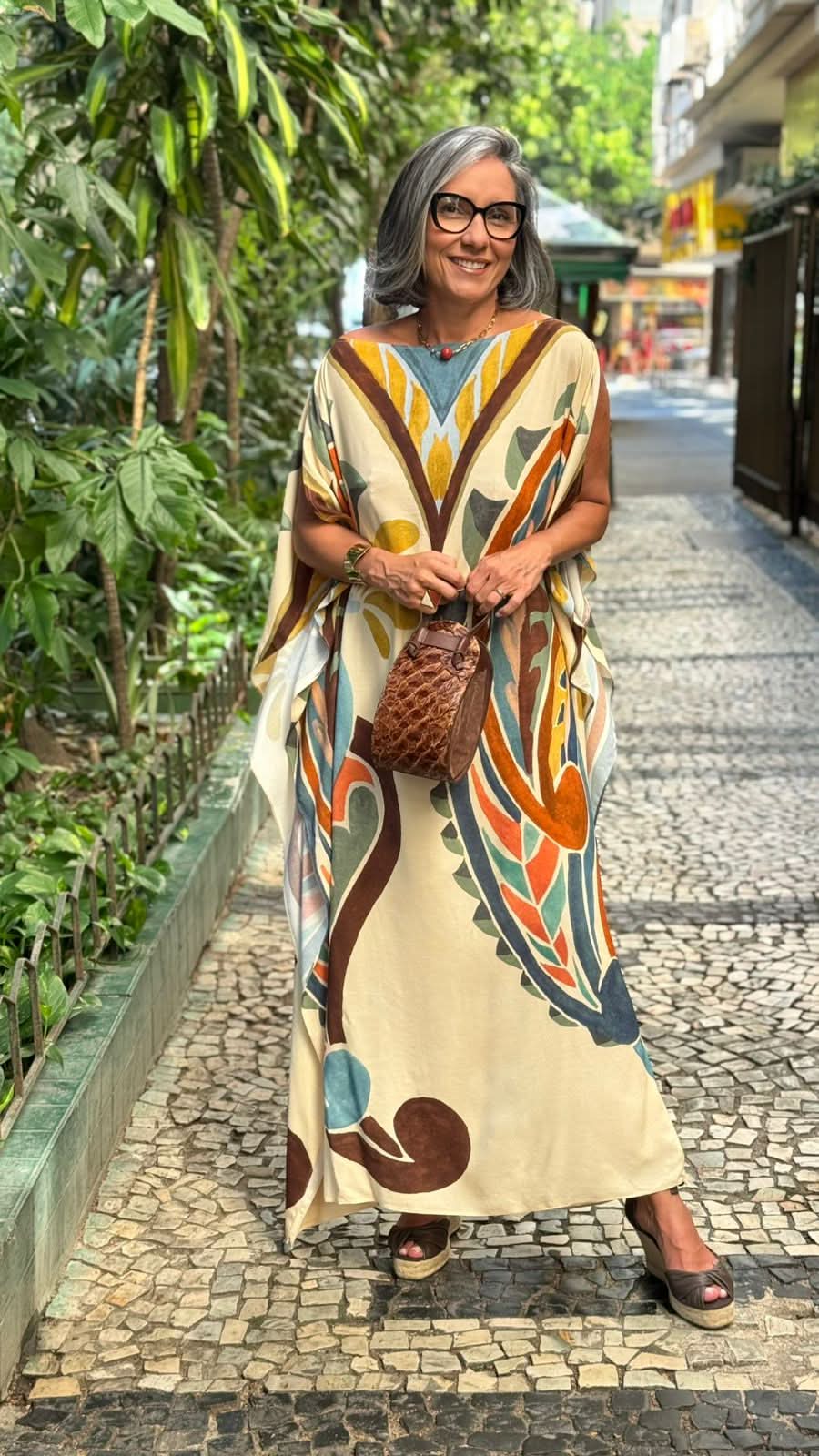 Mulher usando kaftan com estampa colorida, bolsa marrom e sandálias, em calçada urbana arborizada EZILDINHA