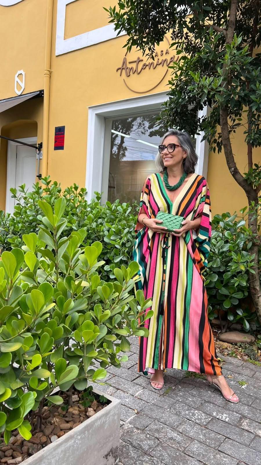 Mulher madura usando kaftan listrado colorido, bolsa verde e colar, em cenário urbano com plantas EZILDINHA