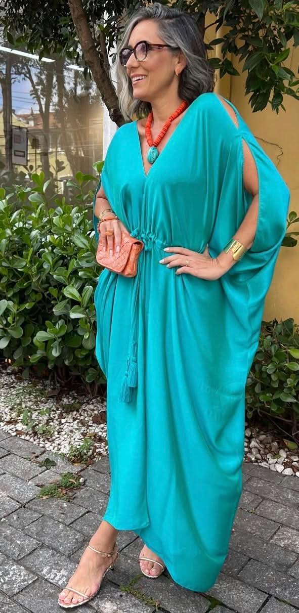 Mulher usando kaftan verde de seda com acessórios coloridos, em ambiente externo com plantas EZILDINHA