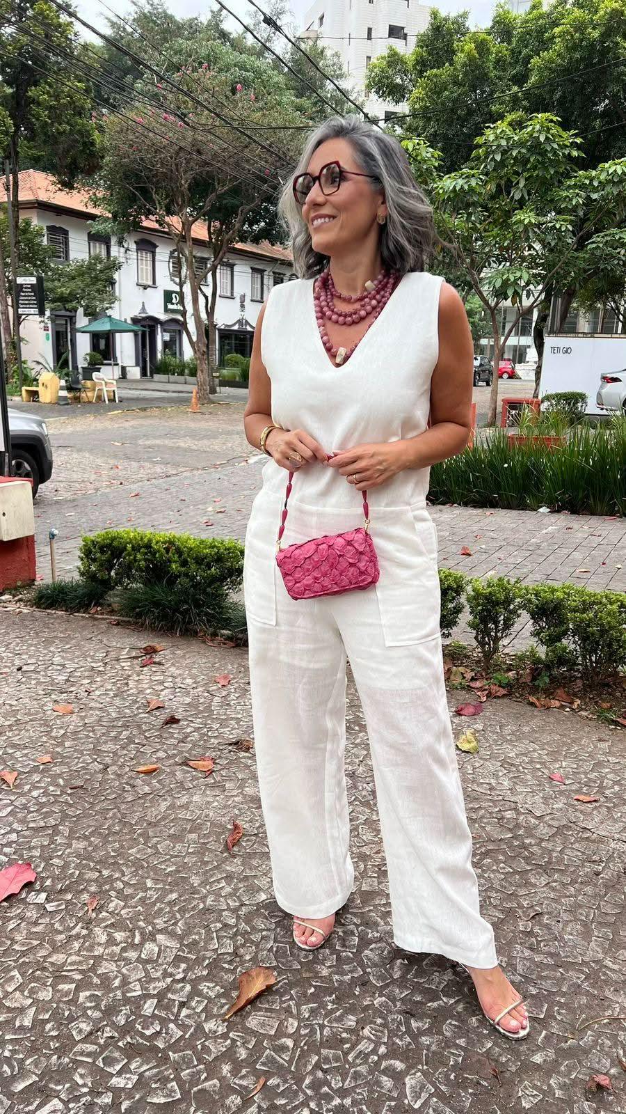 Mulher madura com macacão de linho branco, bolsa rosa, colar e sandálias em rua arborizada EZILDINHA