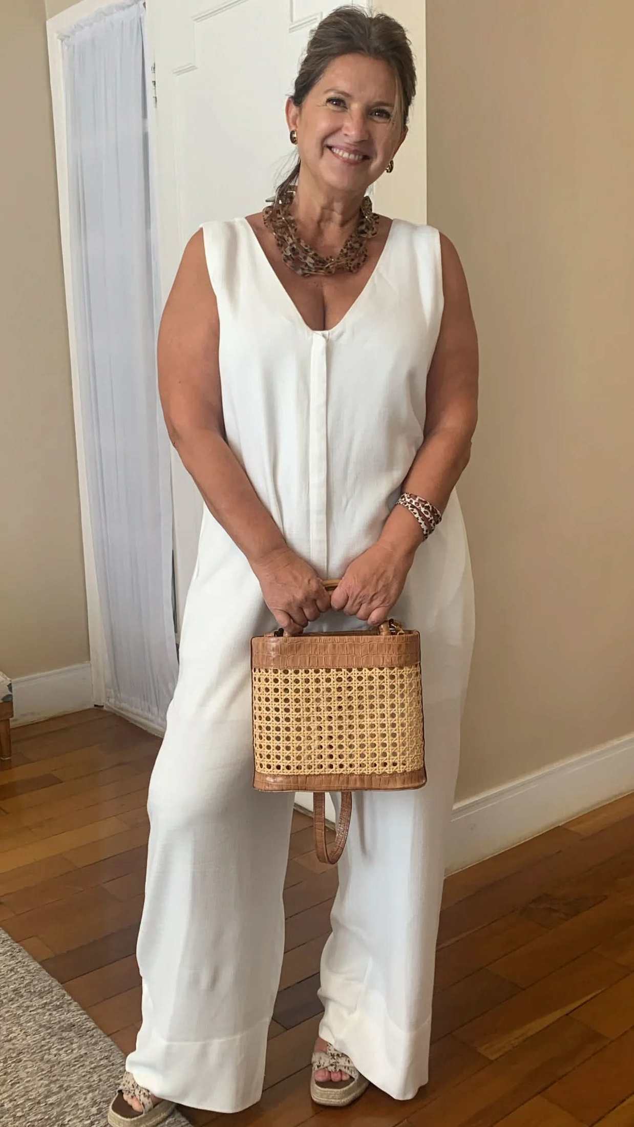 Mulher usando macacão de viscose branco sem mangas, segurando bolsa de palha e sorrindo EZILDINHA