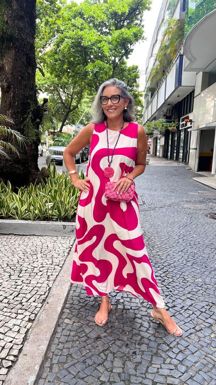 Mulher sorridente usa vestido longo estampado em rosa e branco, sandálias e bolsa rosa na calçada urbana EZILDINHA