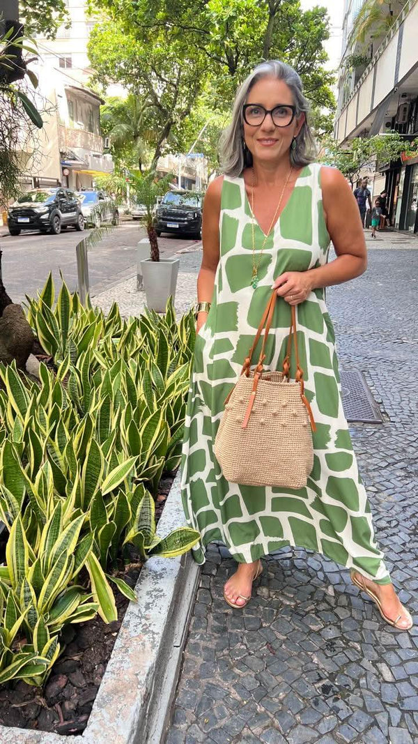 Mulher com vestido longo verde e branco geométrico, bolsa de palha, em calçada urbana arborizada EZILDINHA