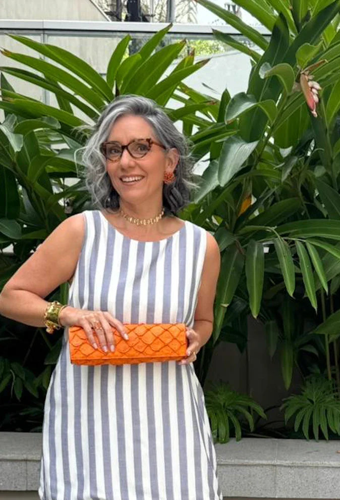 Mulher de cabelos grisalhos usando vestido listrado e segurando clutch laranja, em jardim EZILDINHA