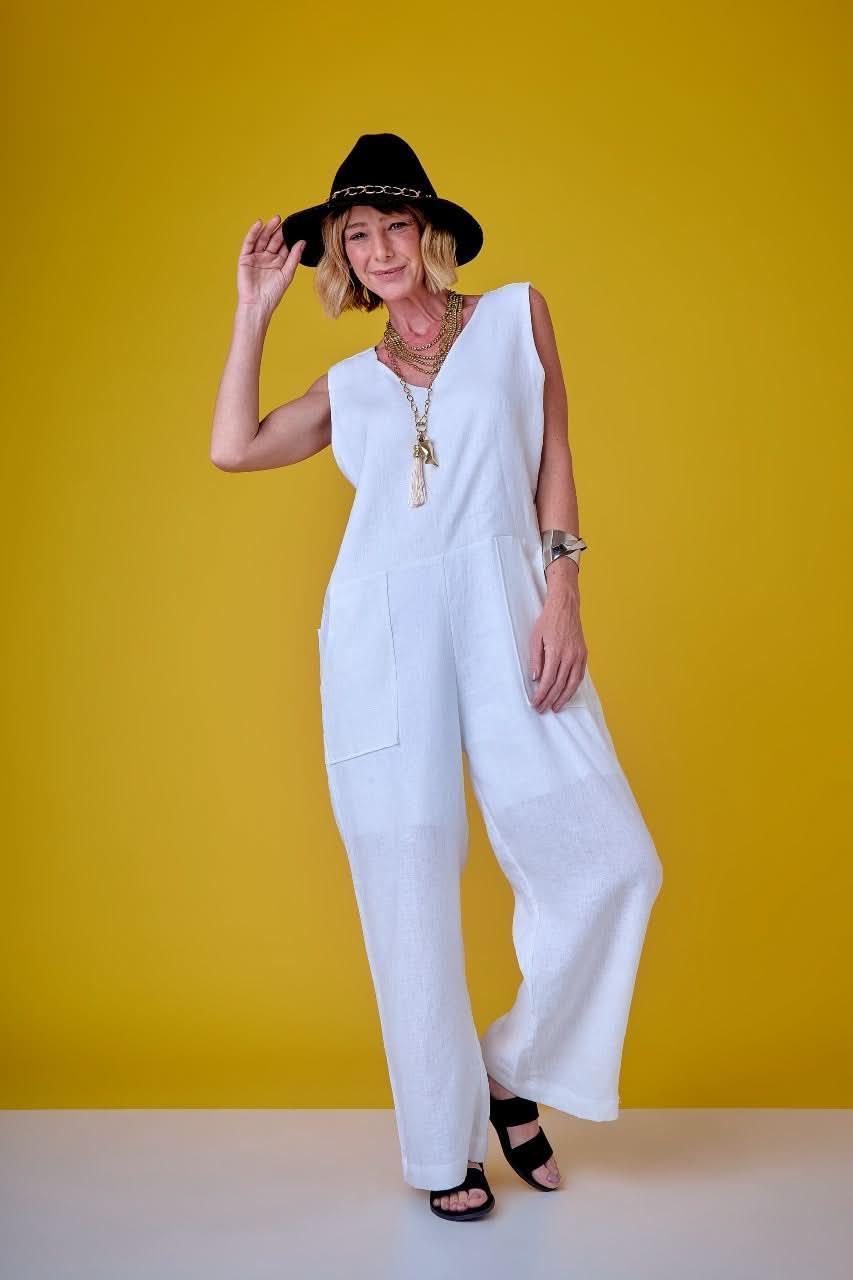 Mulher usando macacão branco de linho, chapéu preto e acessórios dourados, fundo amarelo EZILDINHA
