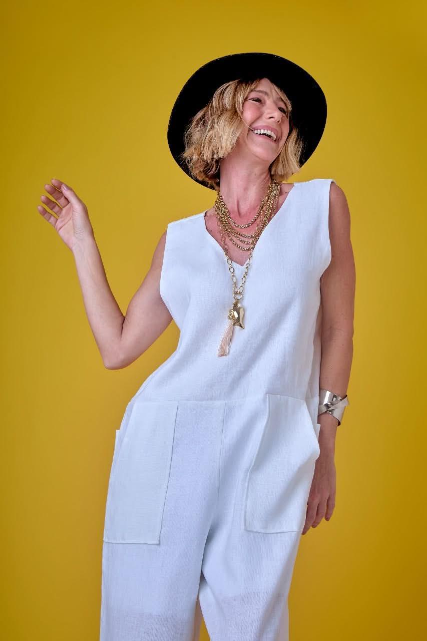 Mulher sorrindo usa macacão branco de linho, chapéu preto e colares, fundo amarelo EZILDINHA