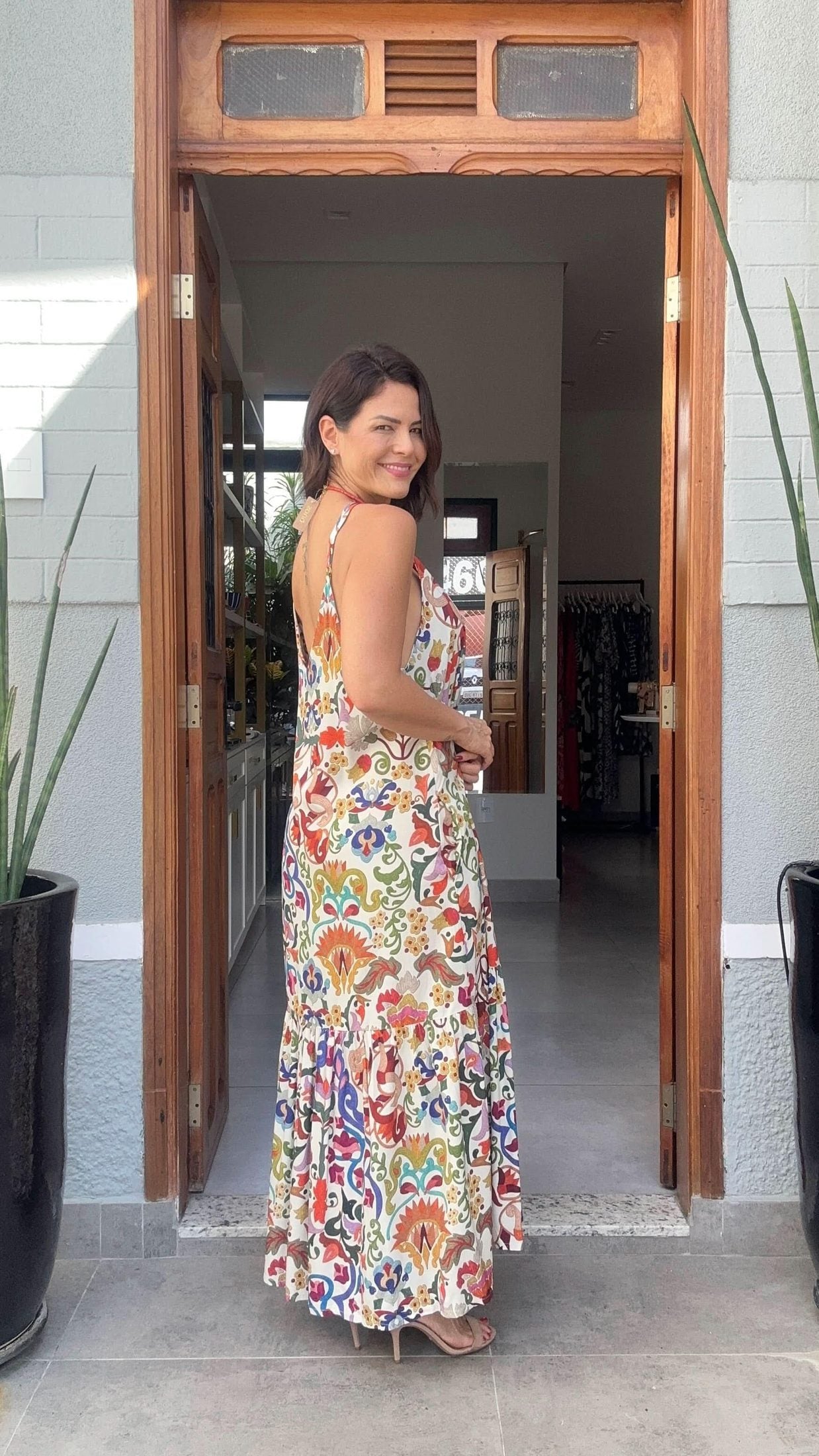 Mulher usando vestido longo de alças com estampa colorida, posando em porta de entrada EZILDINHA