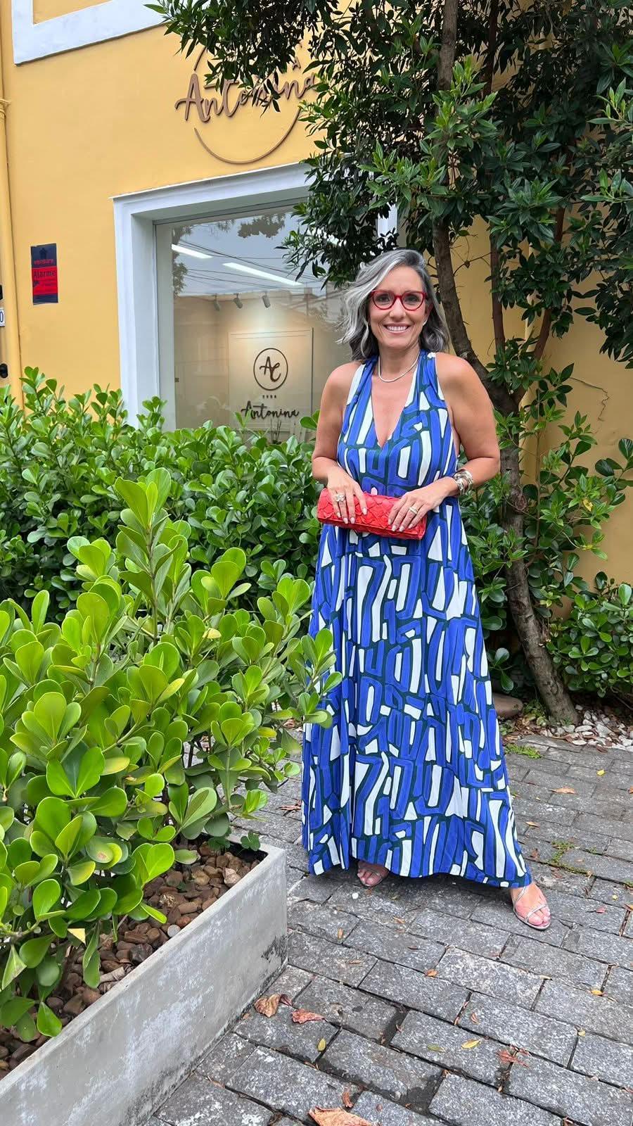 Mulher com vestido longo azul estampado geométrico, segurando clutch vermelha, em área externa com plantas EZILDINHA