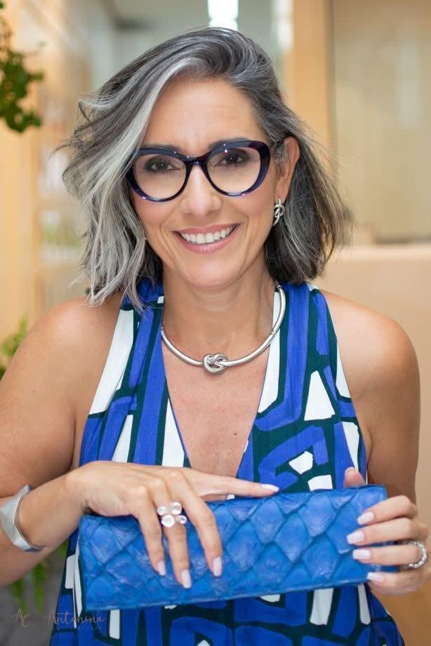 Mulher de cabelos grisalhos usa vestido geométrico azul e segura clutch azul, sorrindo EZILDINHA